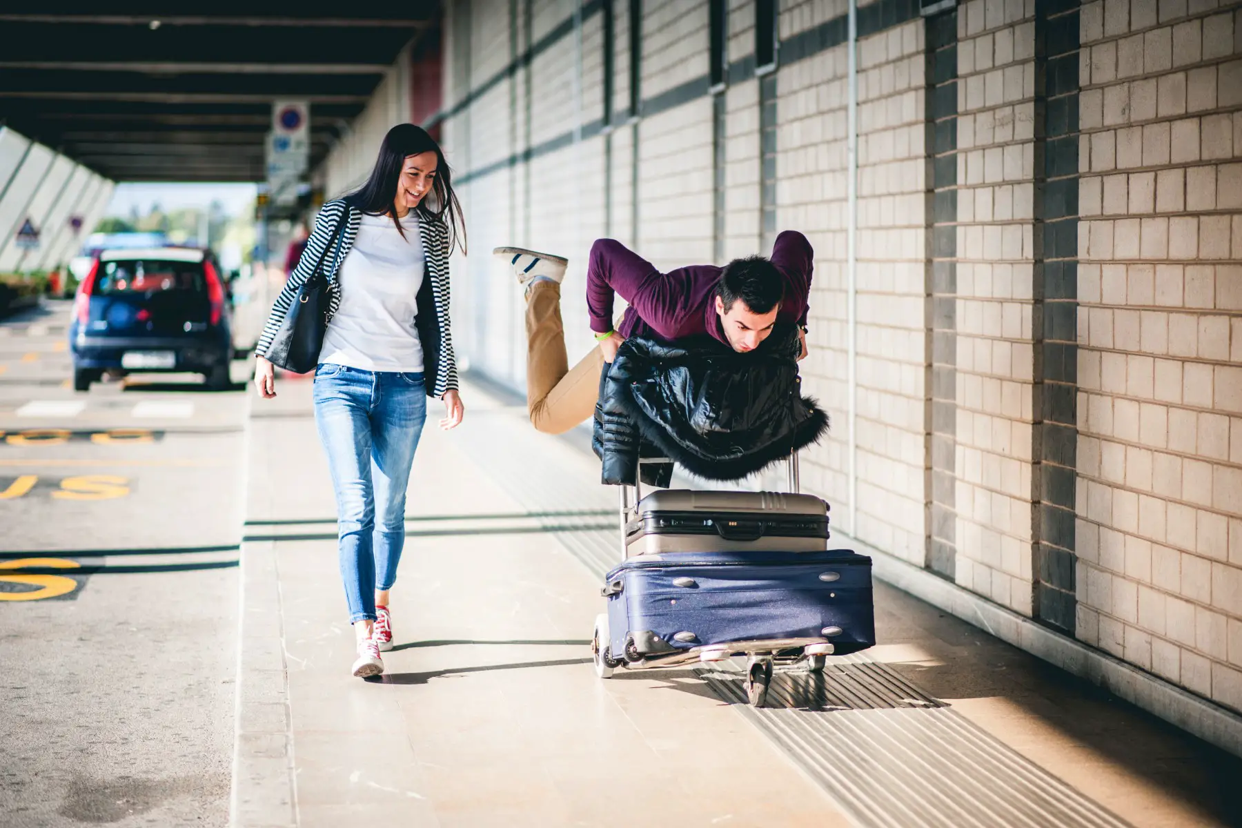 Young couple walking with a trolley and two suitcases at the airport, the woman is laughing while the man is acting silly.