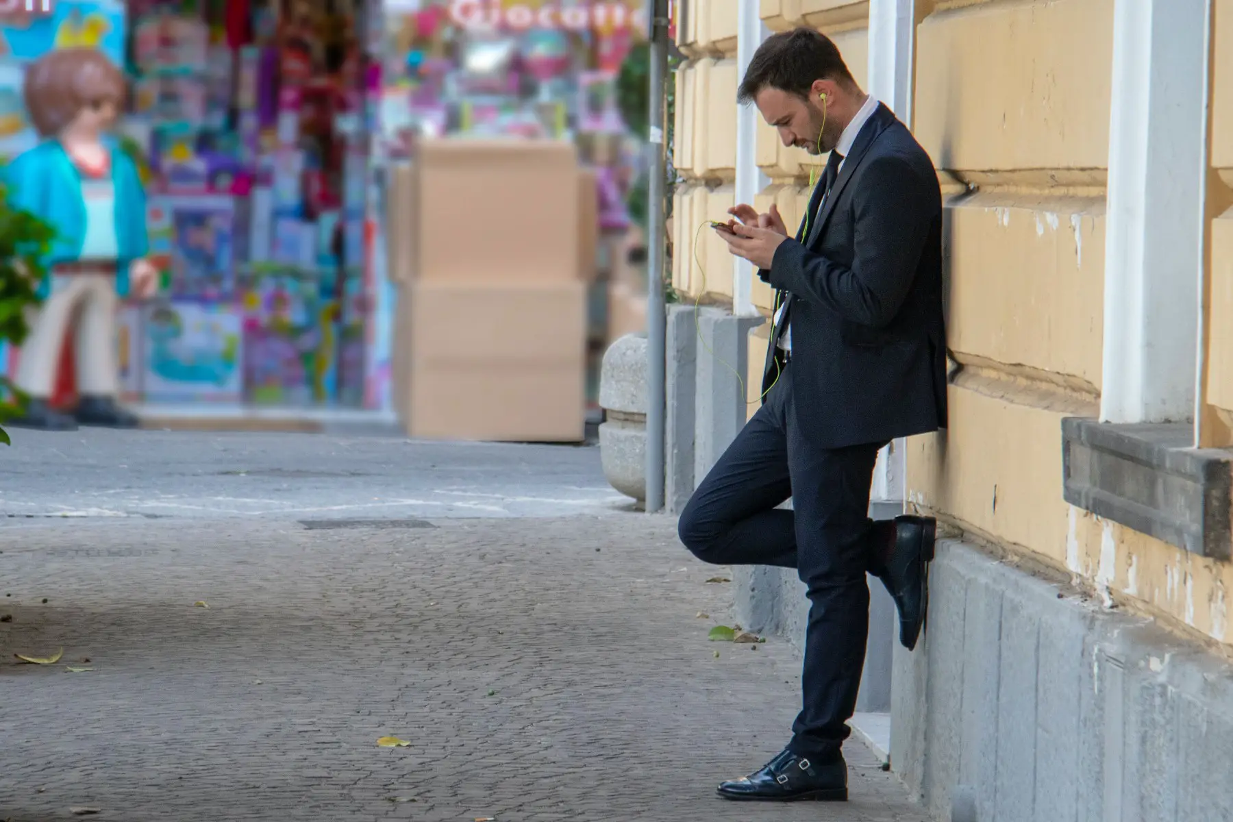 A man in a suit with earbuds in, leans against a wall in Sorrento (Italy), staring intently at his smartphone.
