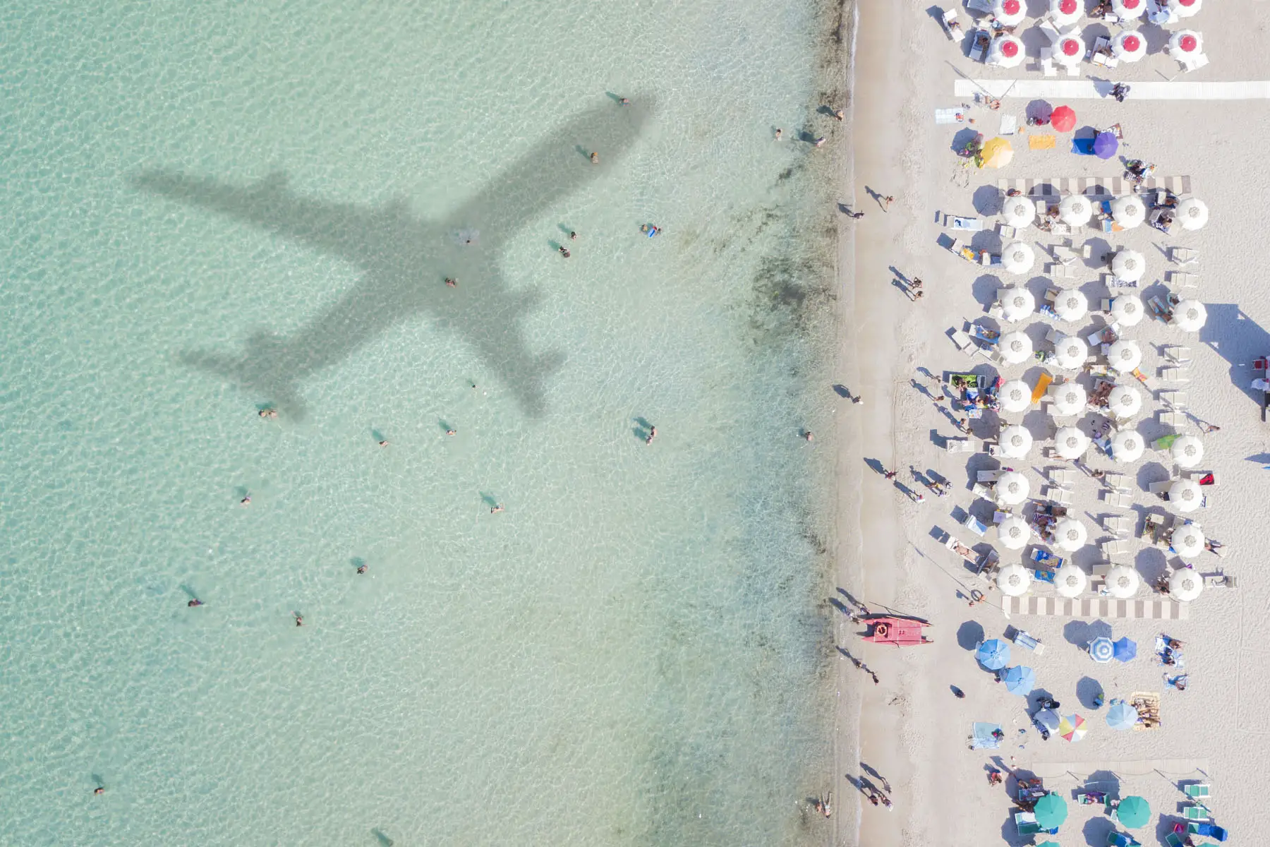 Aerial view of azure beach front with the shadow of an airplane and people who swim.