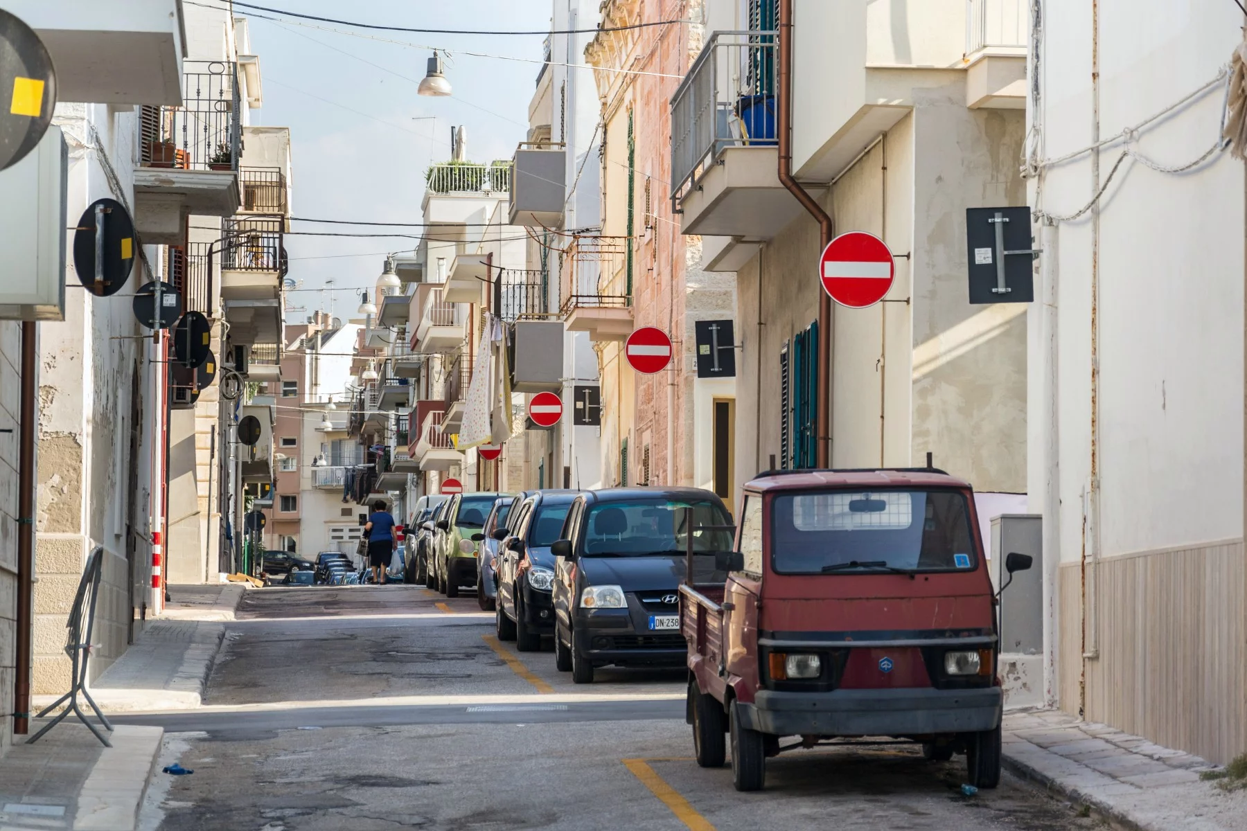 Woman walking along parked cars on a street in Polignano a Mare, Italy, with many no entry traffic signs.