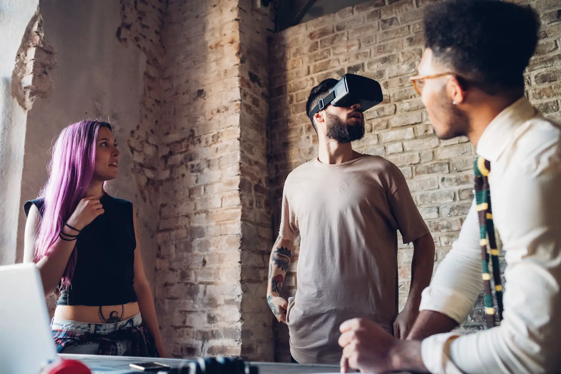 Three people in a loft, one is wearing VR goggles, while the other two are looking at him.