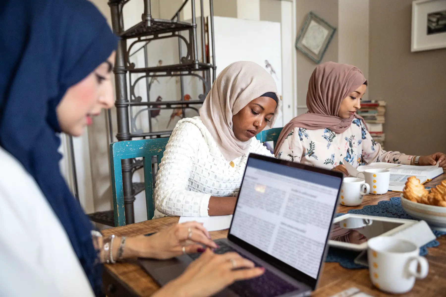Three women with headscarves studying at home, one is working on a laptop.