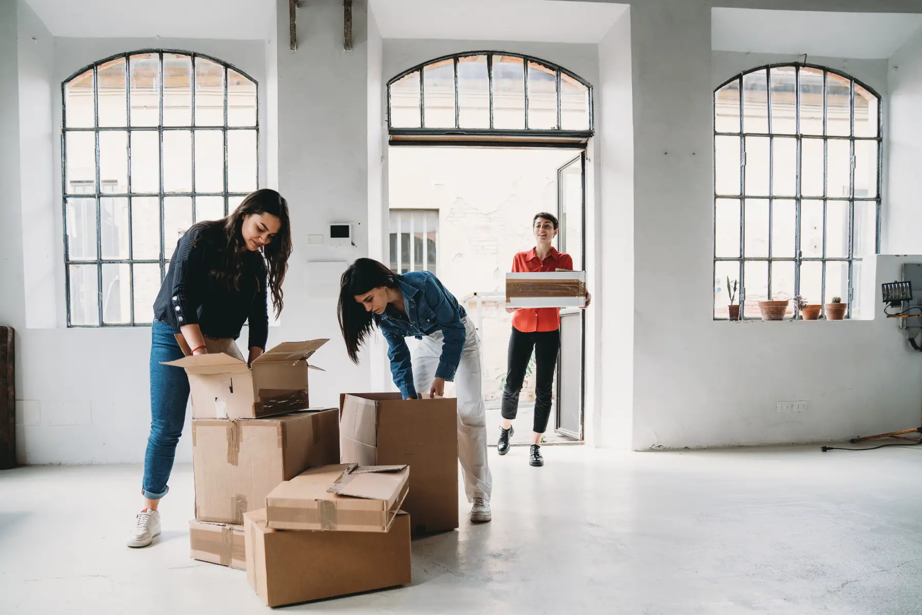 Two women are unpacking boxes in their new loft, a third one is entering the apartment carrying another box.