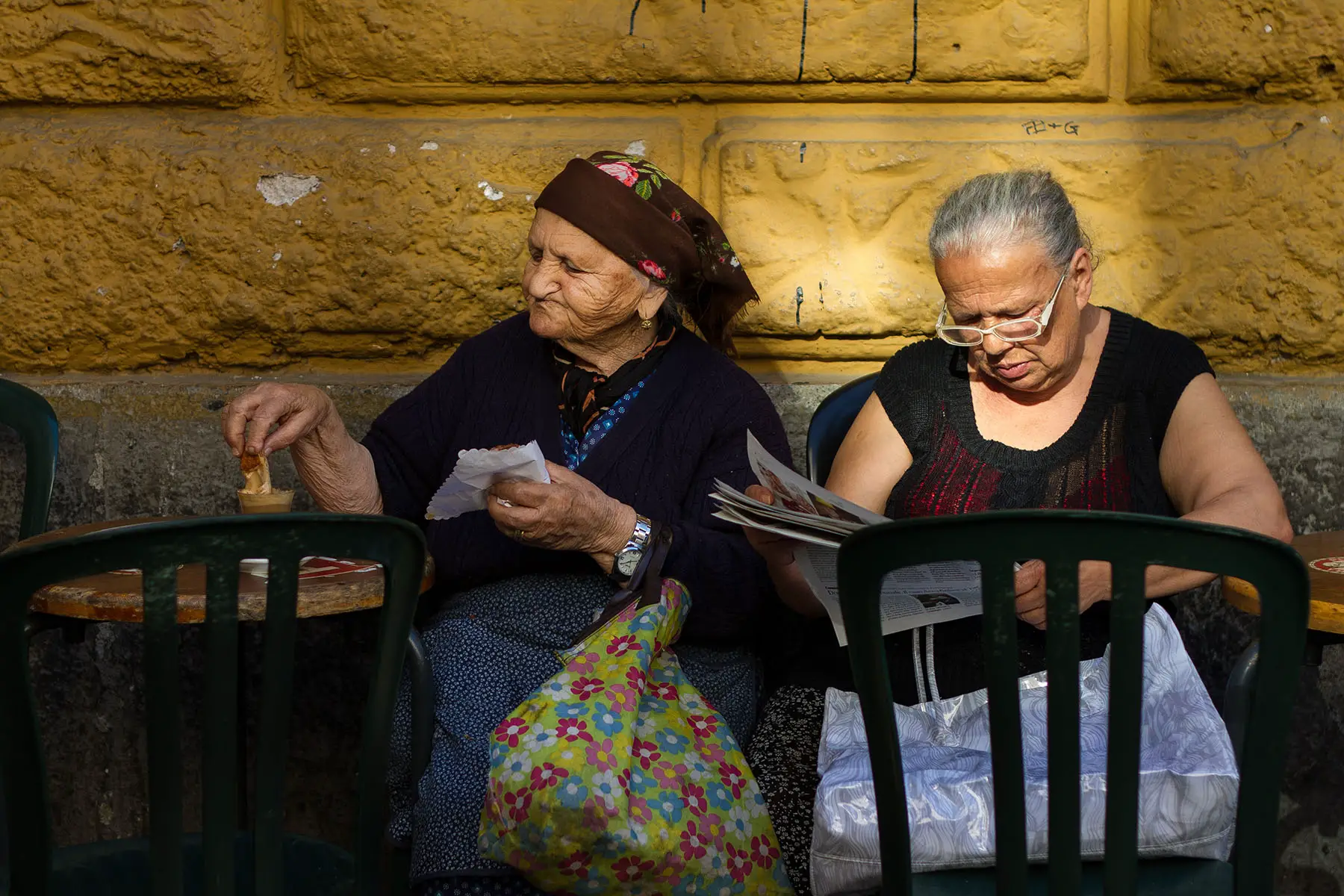 Two senior women relax against a sunlit yellow wall at an outdoor cafe in early morning in the Trastevere neighborhood. One woman reads the paper while the other dunks a pastry into her coffee.