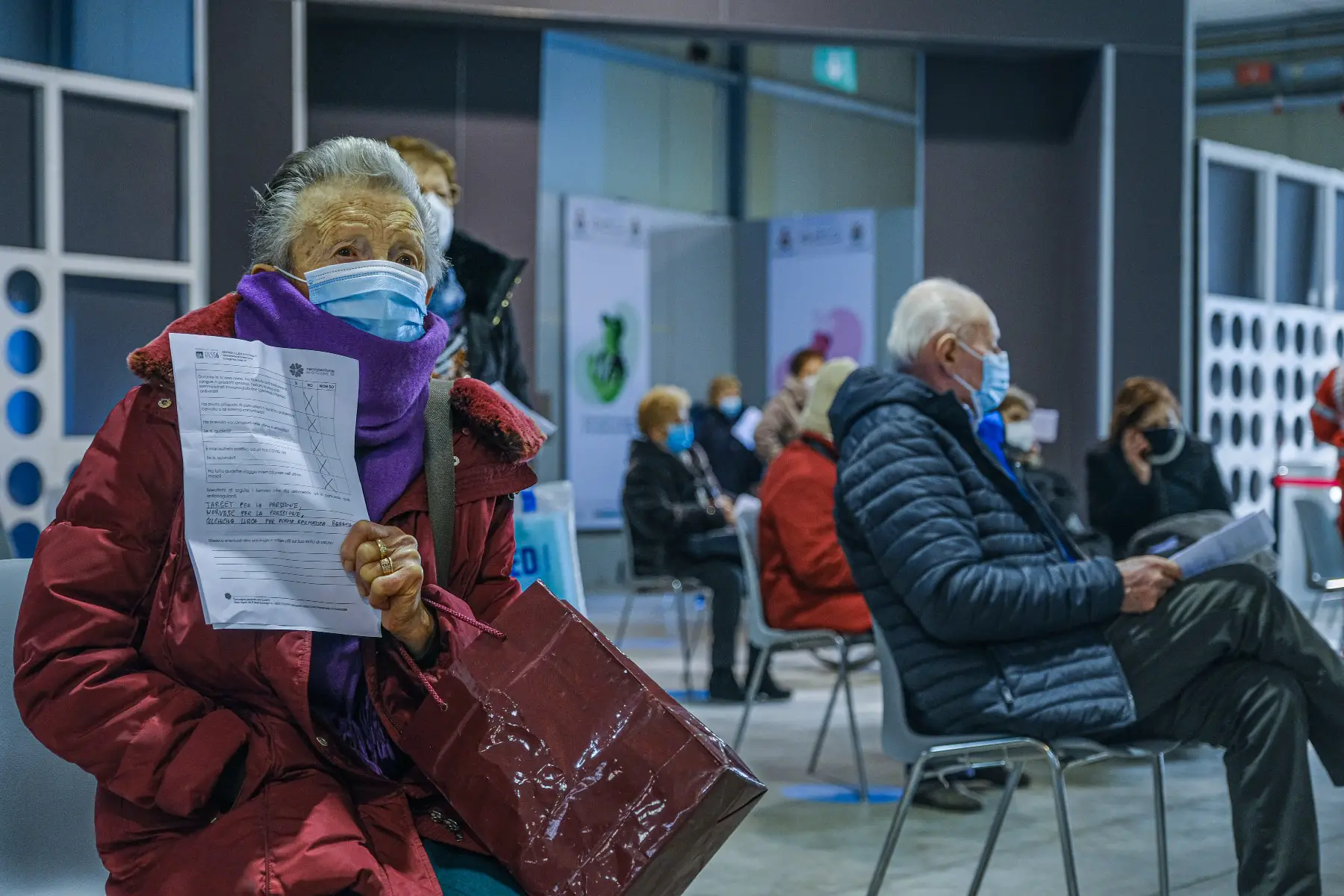 Mature adults waiting to get the COVID-19 vaccine in Padua, Italy. All are wearing protective face masks and are sitting three feet apart. An older woman is sitting in the foreground, hand clutching a filled out questionnaire.