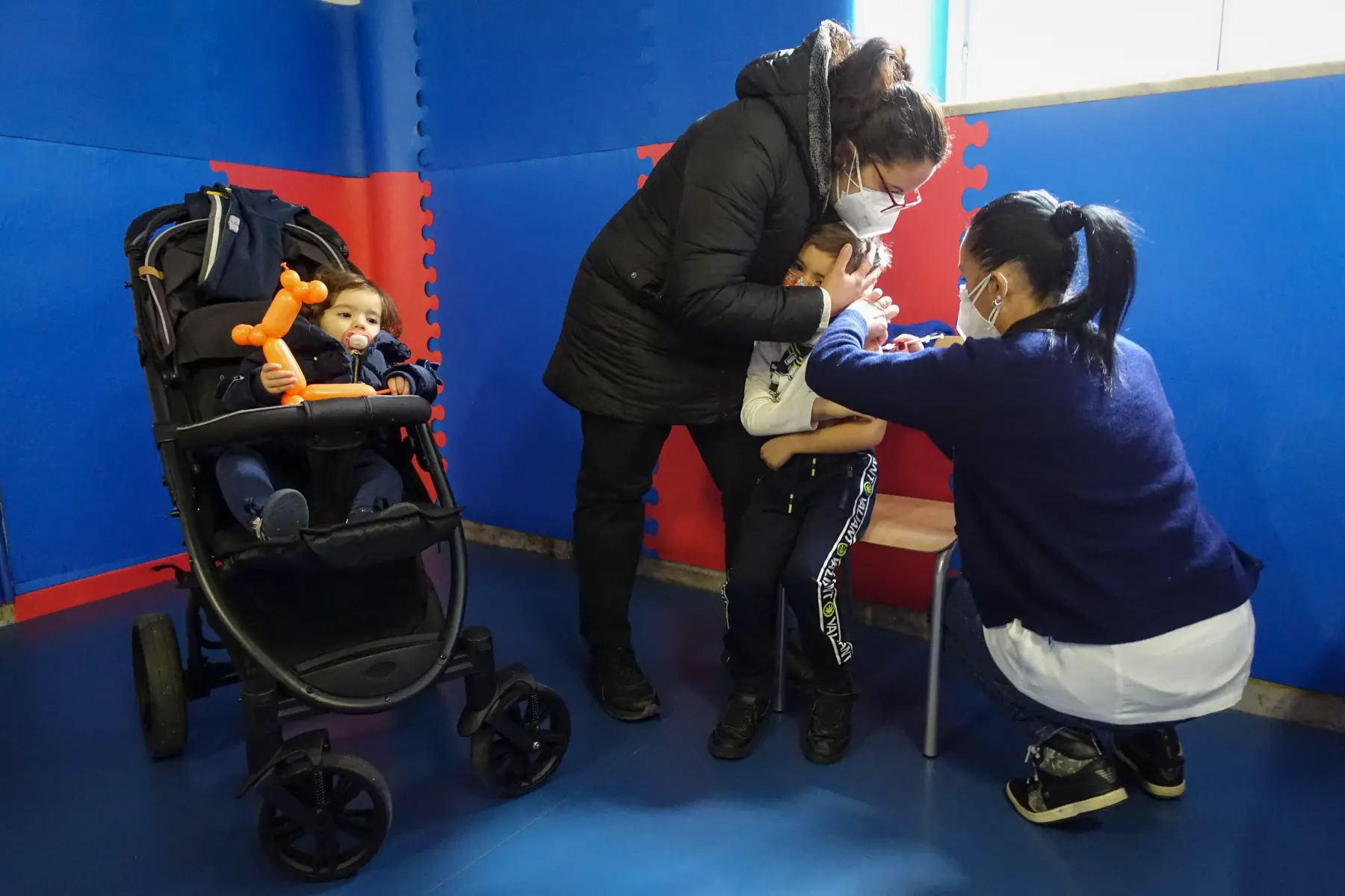 Healthcare professional is administering a COVID vaccine to a child, who is held in their mother's arms. Their sister are sitting nearby in a stroller, holding a balloon animal and looking a bit bored.