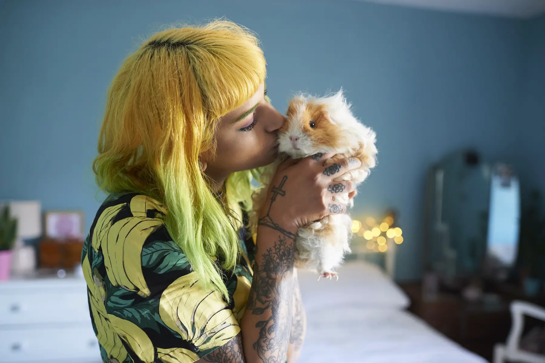 A woman with tattoos and dyed hair holds up her fluffy white and orange guinea pig to kiss its cheek.