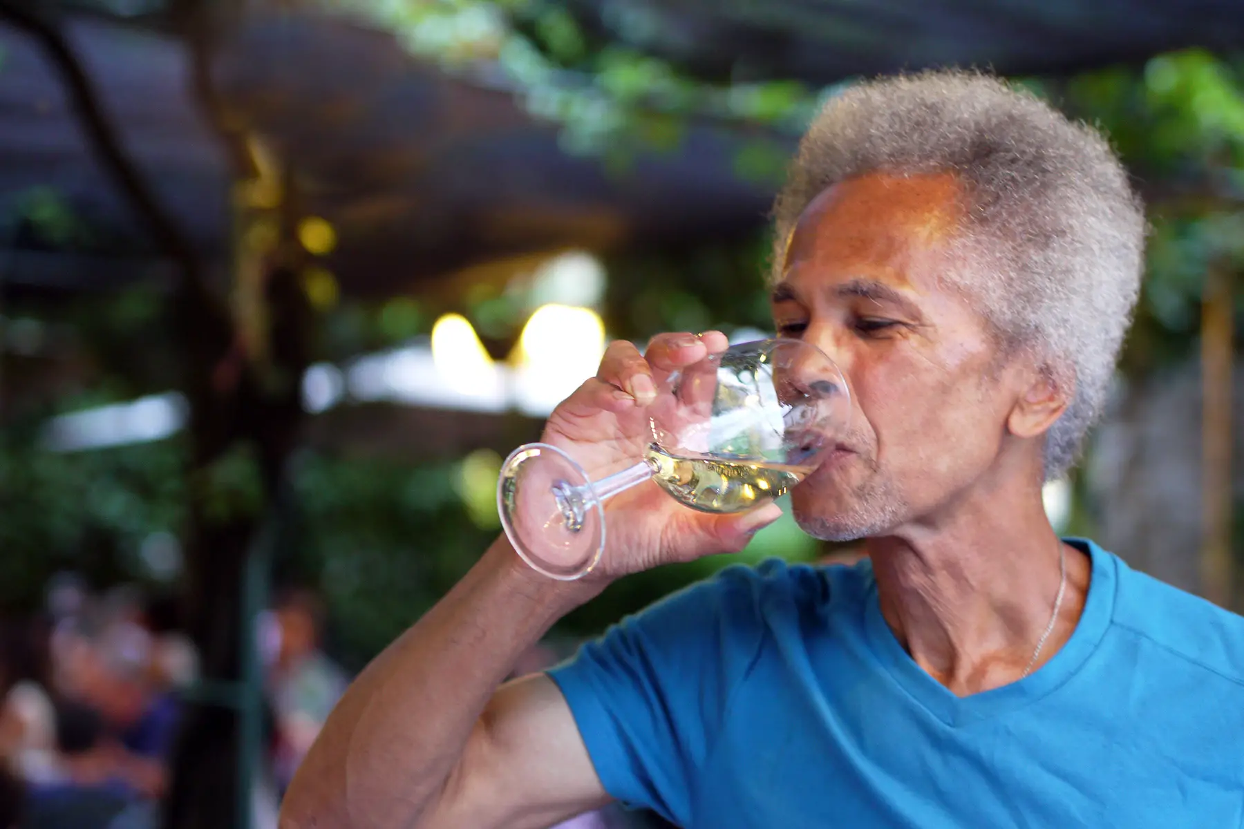 A man with grey hair sipping a white wine and looking happy
