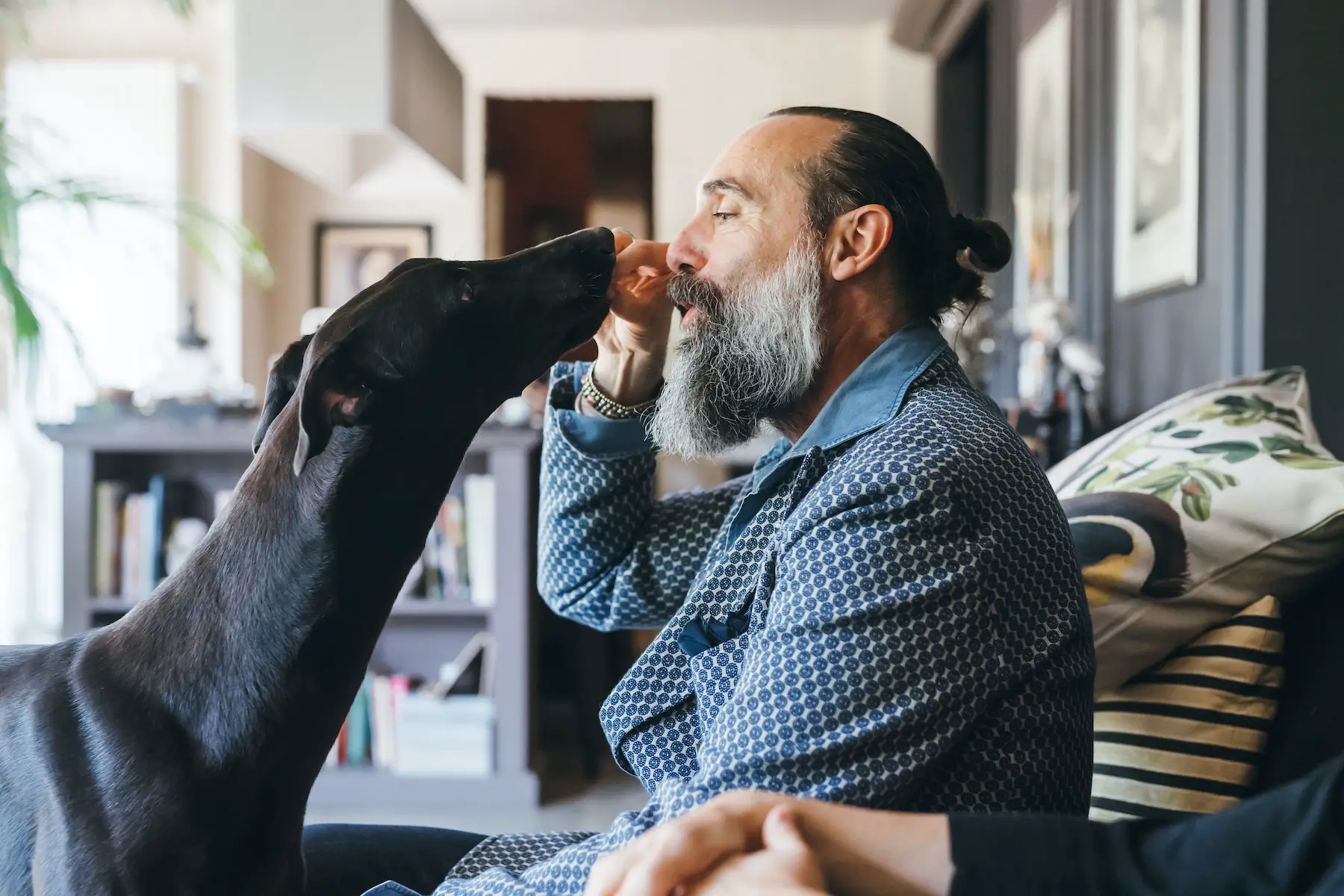 A bearded man holds up a treat for his large black dog to take from right in front of his face.