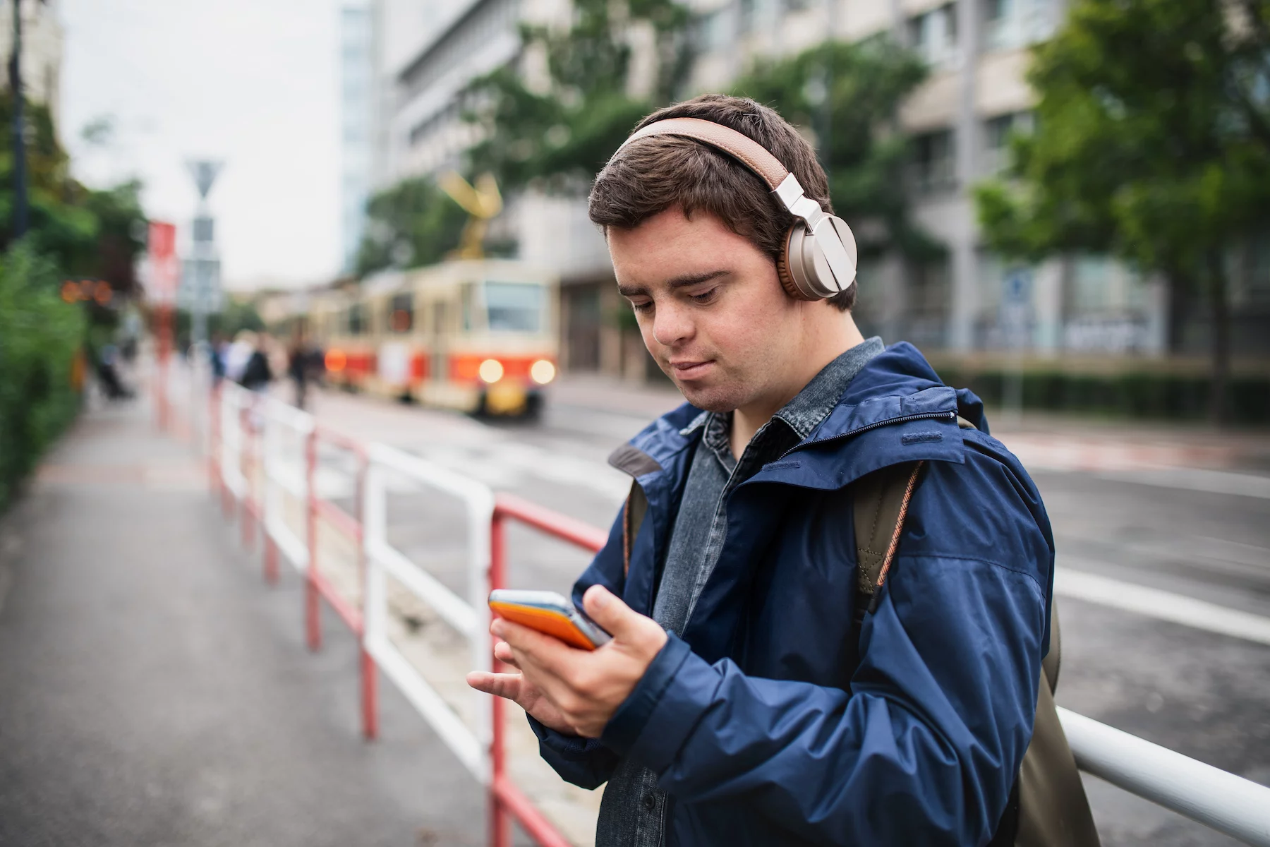 A young man wearing over-ear headphones checks his phone while a tram drives by behind him