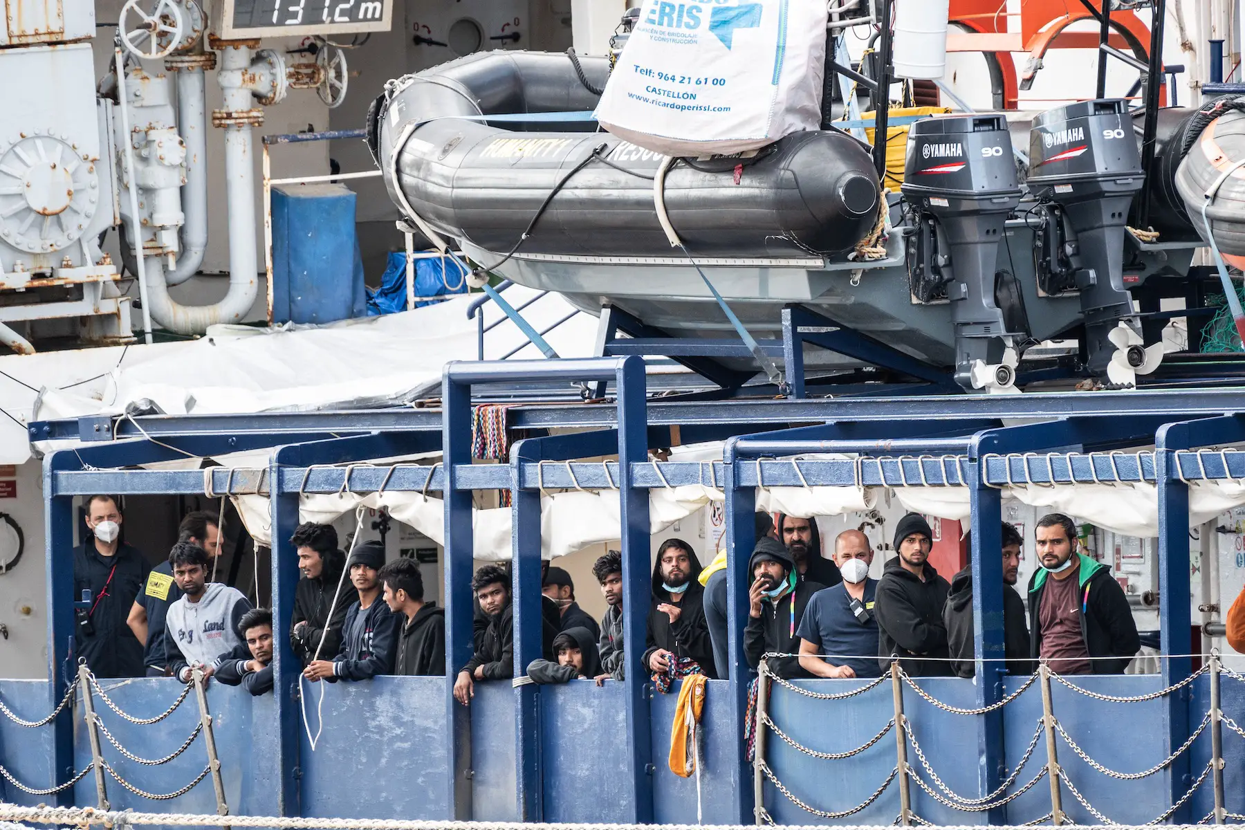 A group of migrant men look out over the edge of a boat docked in Italy's harbor while NGO staff stand masked beside them