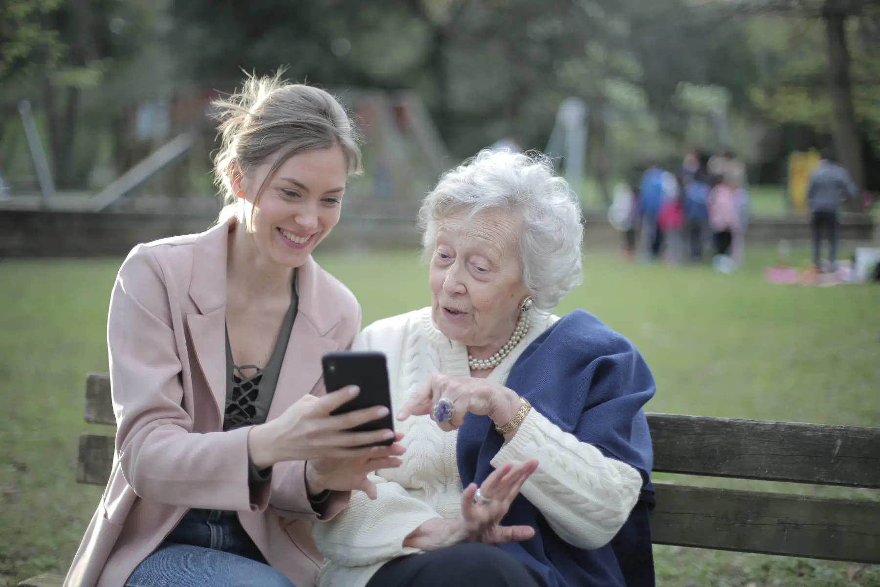Adult daughter showing mother or grandmother how to order groceries using mobile phone apps. Sitting on a park bench.