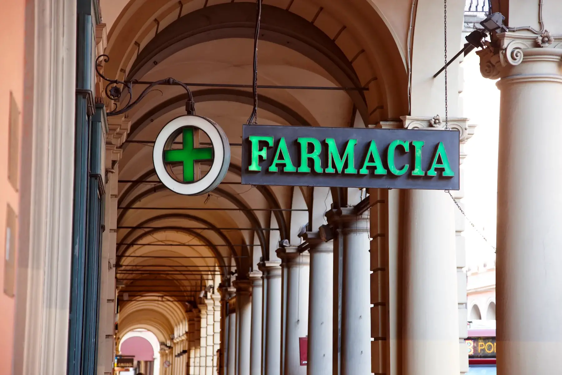 A sign reading pharmacy in Italian (farmacia) hangs from an arched walkway in Bologna