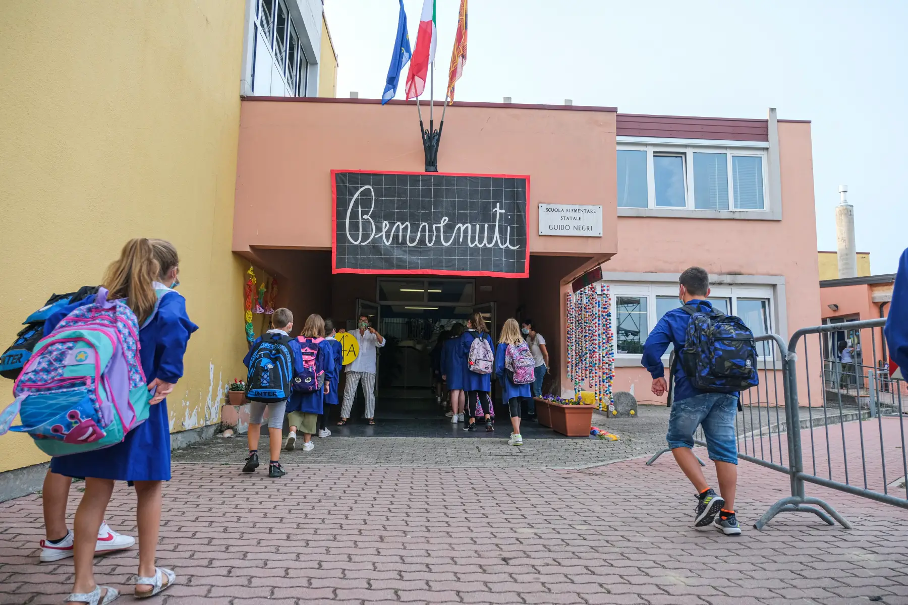 Primary students with backpacks enter their school, a welcome sign reading Benvenuti is above the entrance