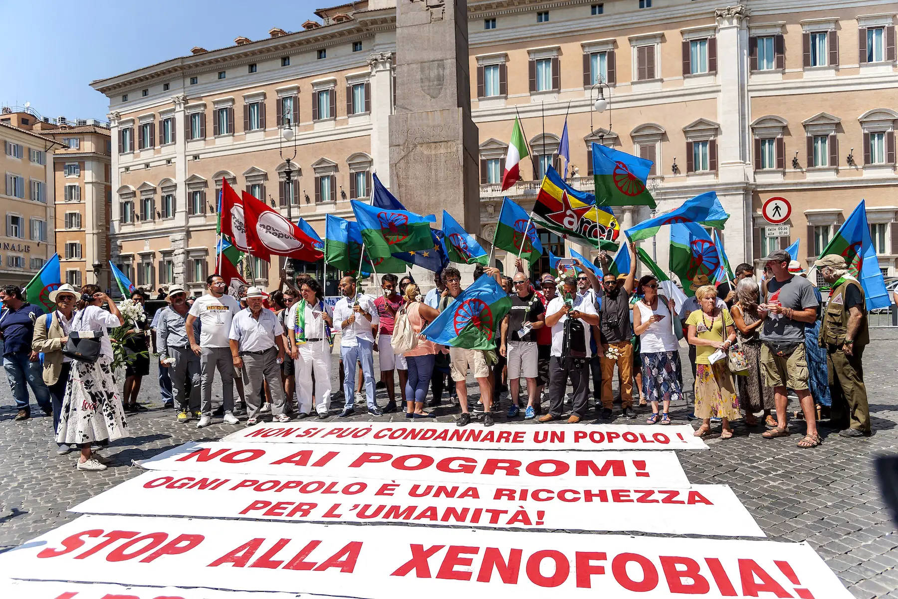 A large gathering of Romani people and supporters fly the Romani flag and display signs on the ground protesting xenophobia