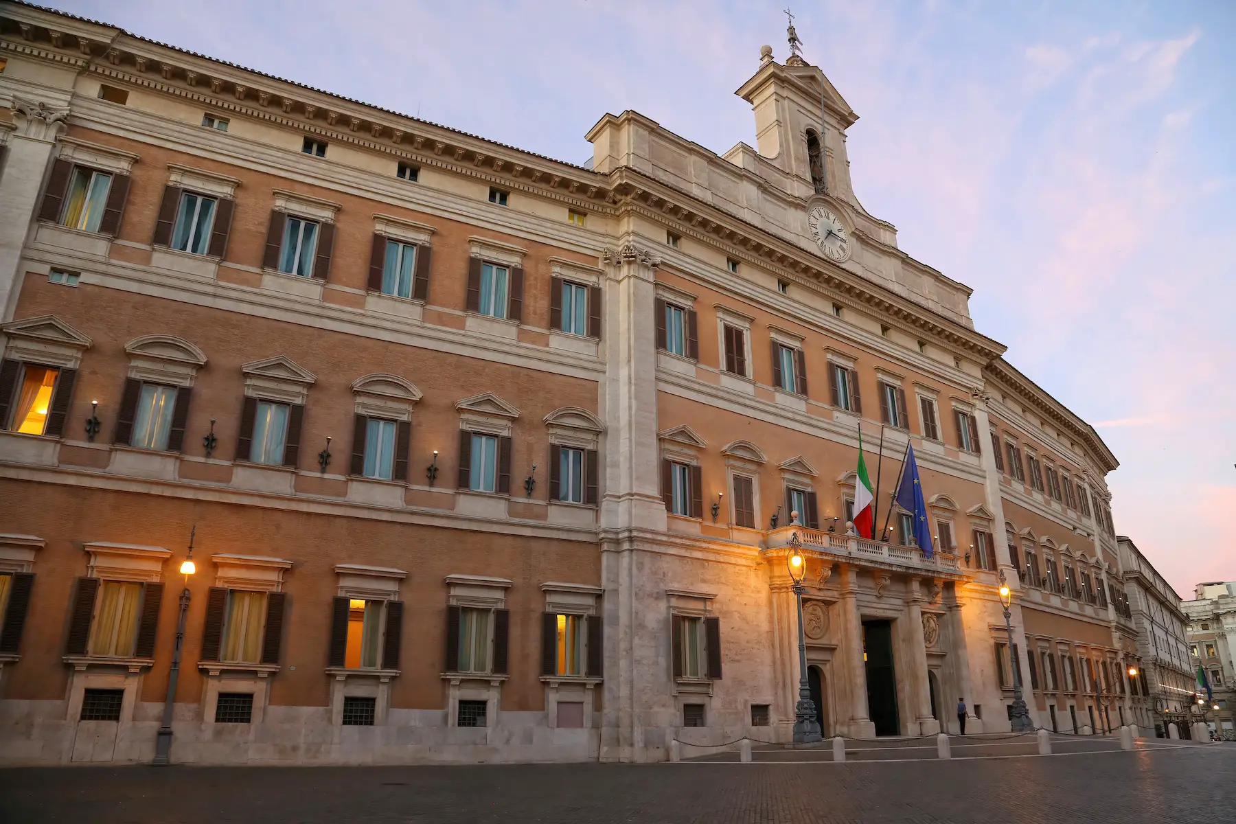 The Italian parliament building in Rome is shown at sunset, an Italian and European Union flag each displayed over the main entrance