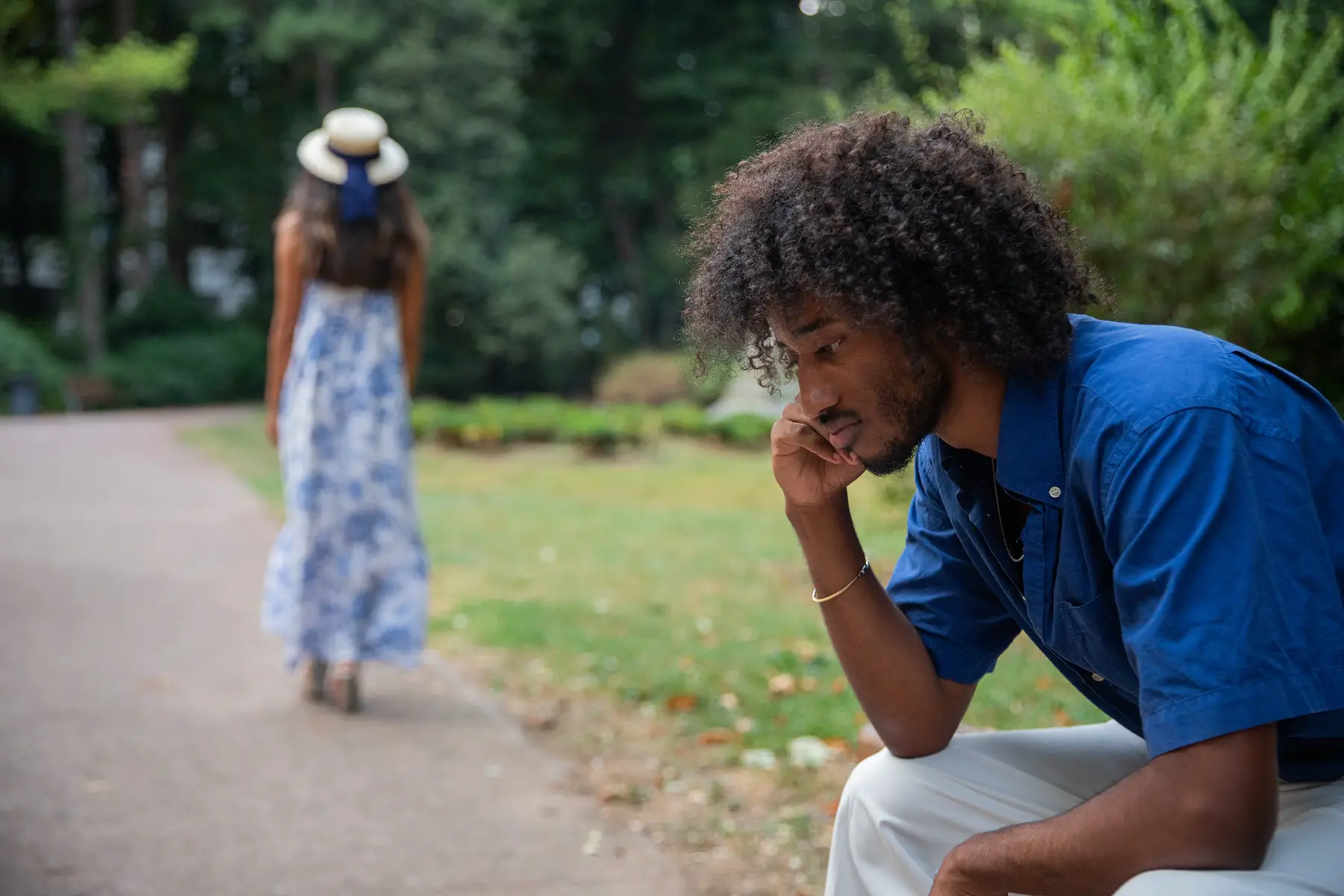 A sad man sits on a bench in the foreground. Out of focus, in the background, a woman in a long dress and hat walks away.