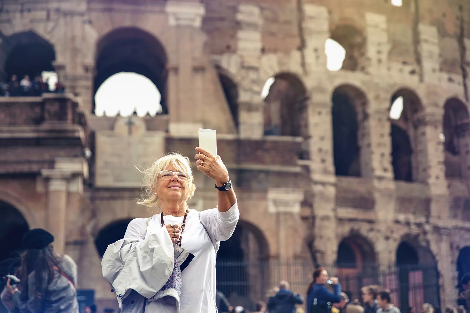 A senior woman takes a selfie while standing front of the colosseum in rome, joys of retiring in Italy