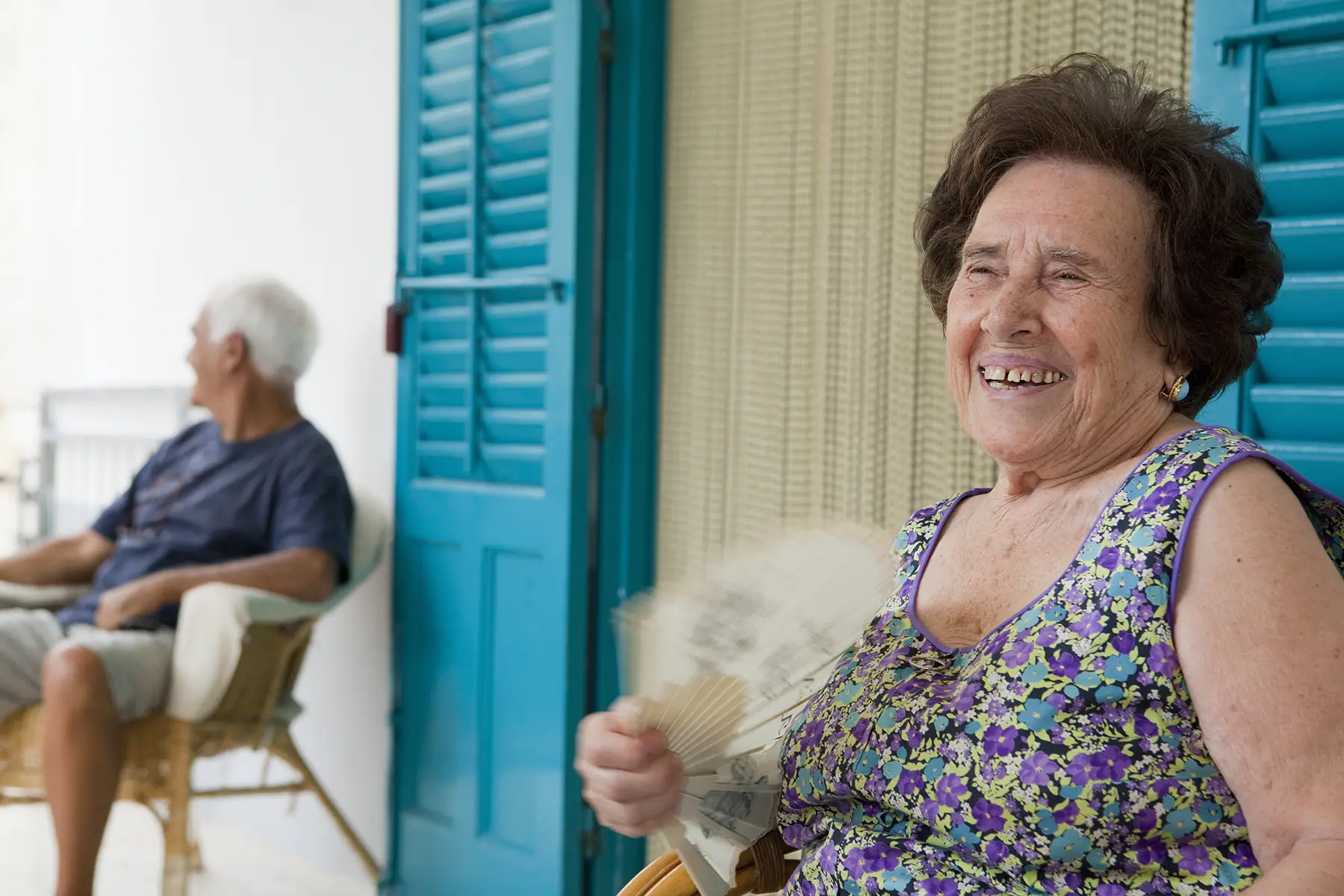 Older woman, laughing and fanning herself, older man sitting in background outside