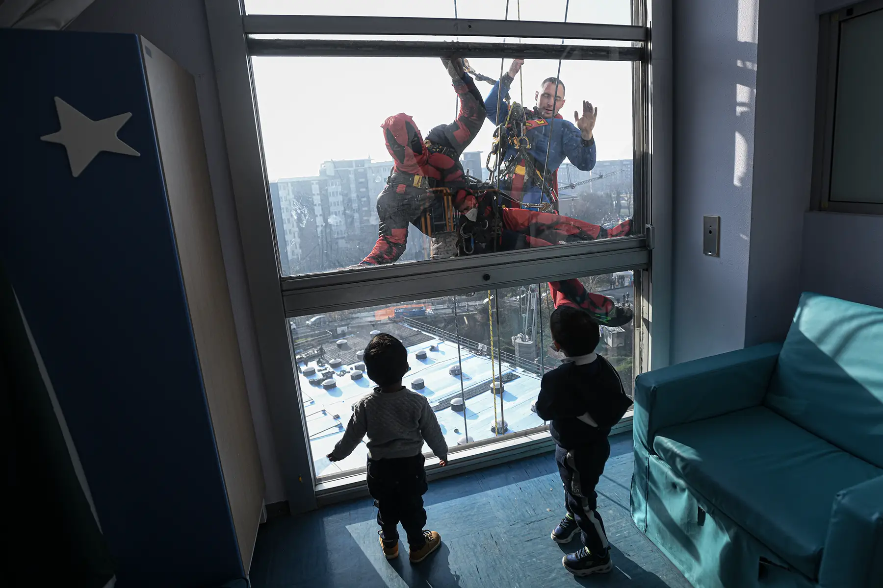 People dressed as superheroes clean the windows of a hospital as two children inside watch them.
