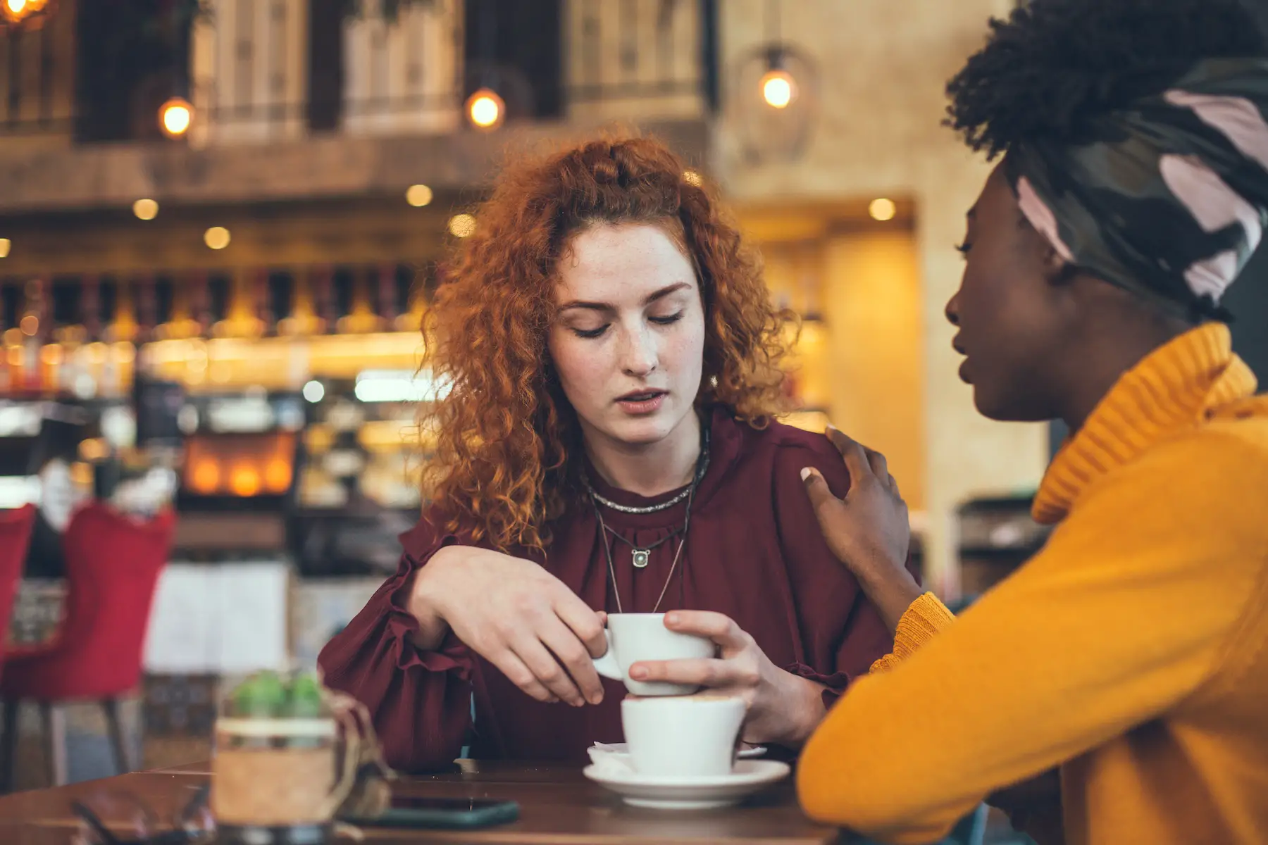 Two women meet for coffee at a cafe in Italy; one looks upset while the other places a hand on her shoulder