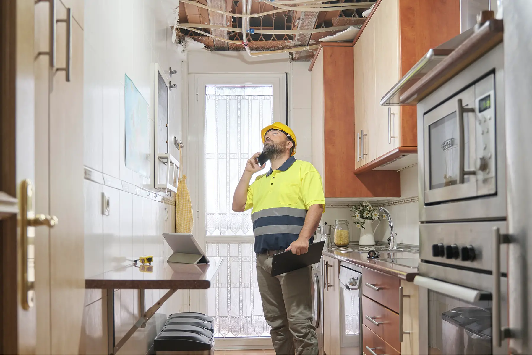A surveyor stands in a kitchen on the phone, looking up at exposed pipes running along the ceiling