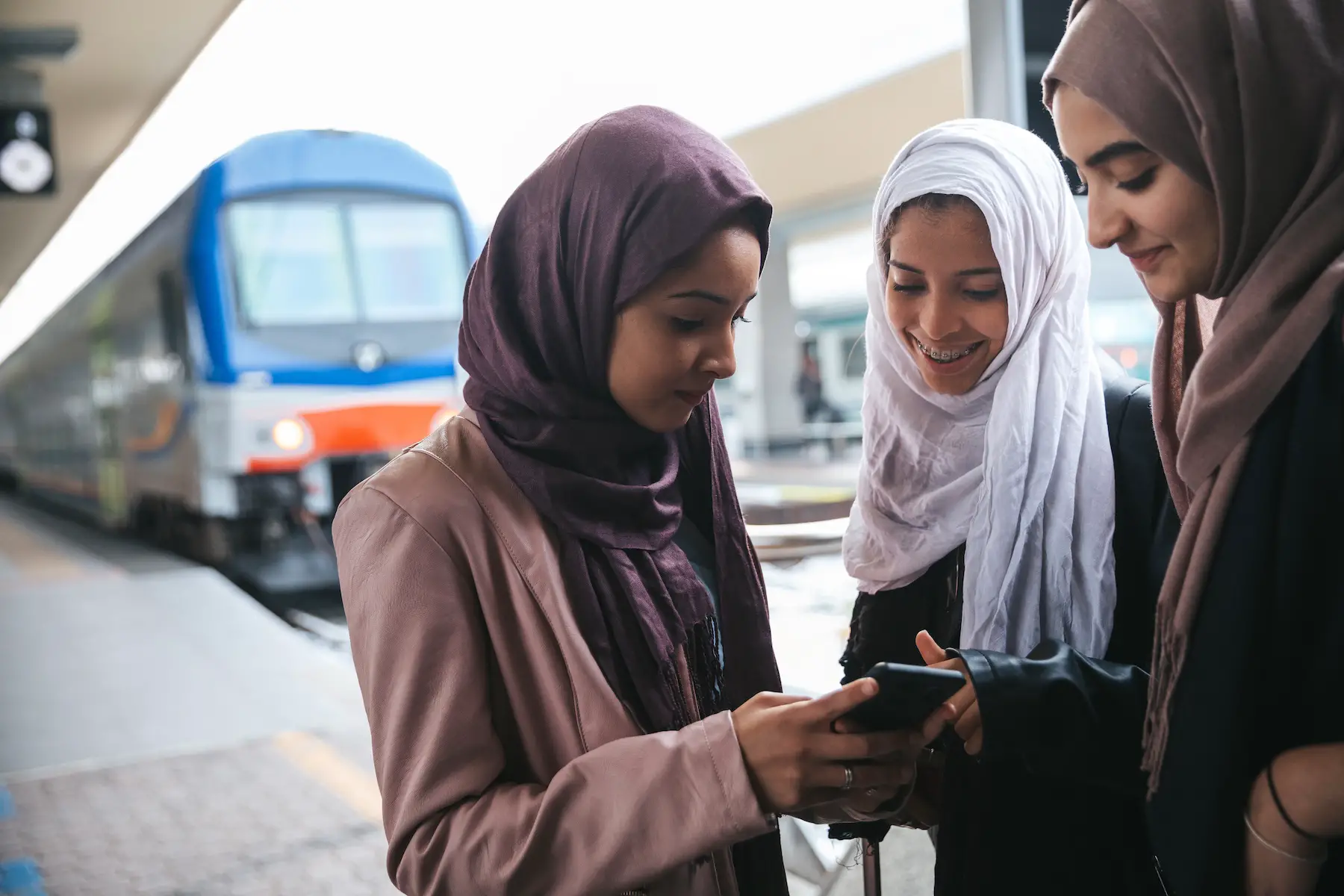 Three young women stand looking at a phone while waiting for the train on the platform