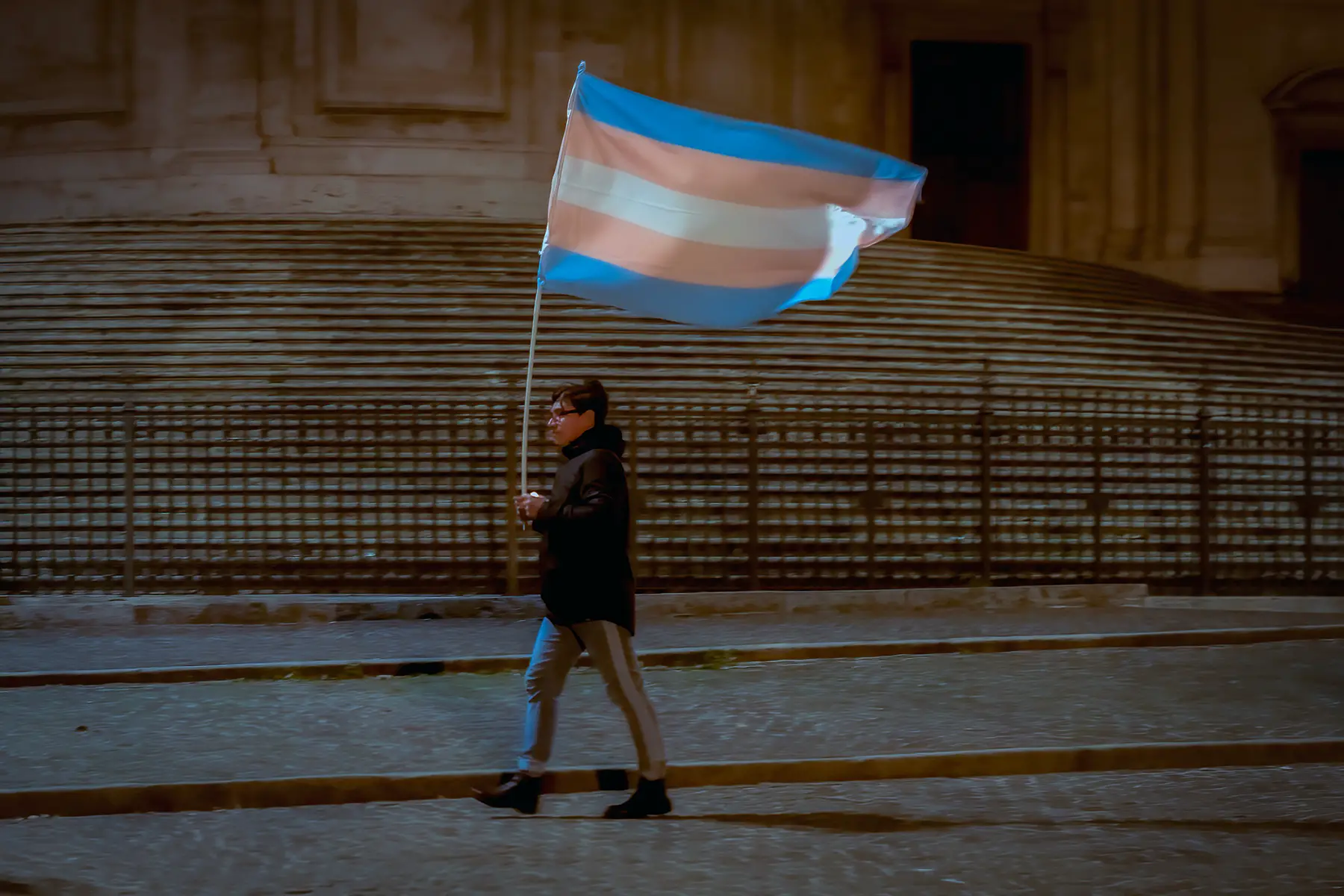 A person walking with a trans flag at night. The trans flag is a horizontal white stripe bordered by two light-pink stripes, which are bordered by two blue stripes)