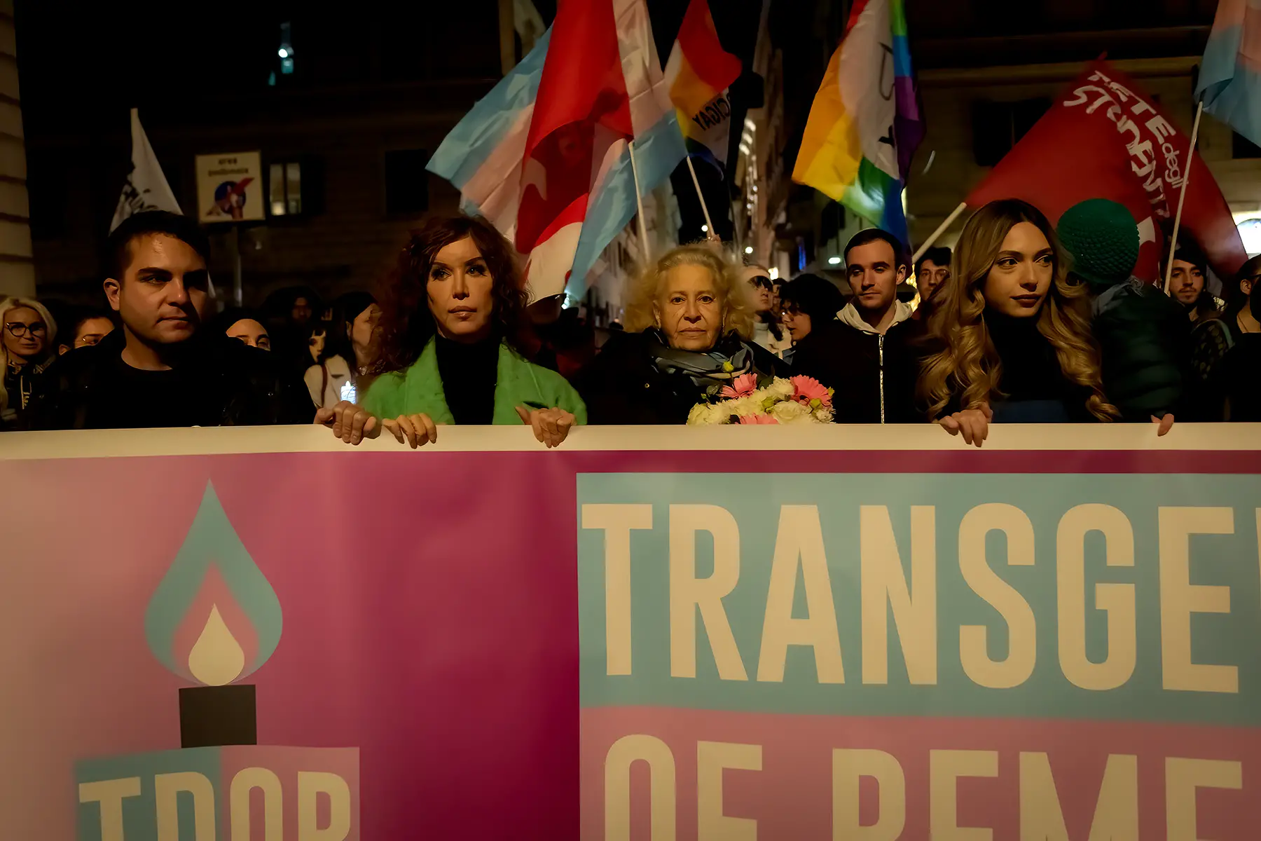 A group of people march with flags and a banner for Transgender Day of Remembrance