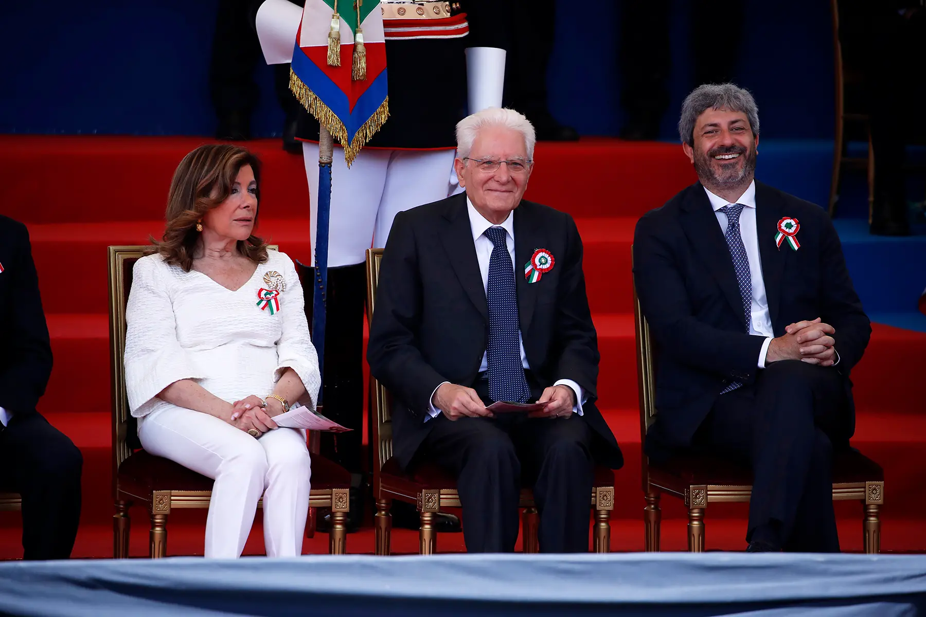 Former President of the Italian Senate Maria Elisabetta Casellati, President of the Republic Sergio Mattarella, and President of the Chamber of Deputies Roberto Fico attending the military parade on the Via dei Fori Imperiali in 2022. All three have small rosettes with the Italian flag on them.