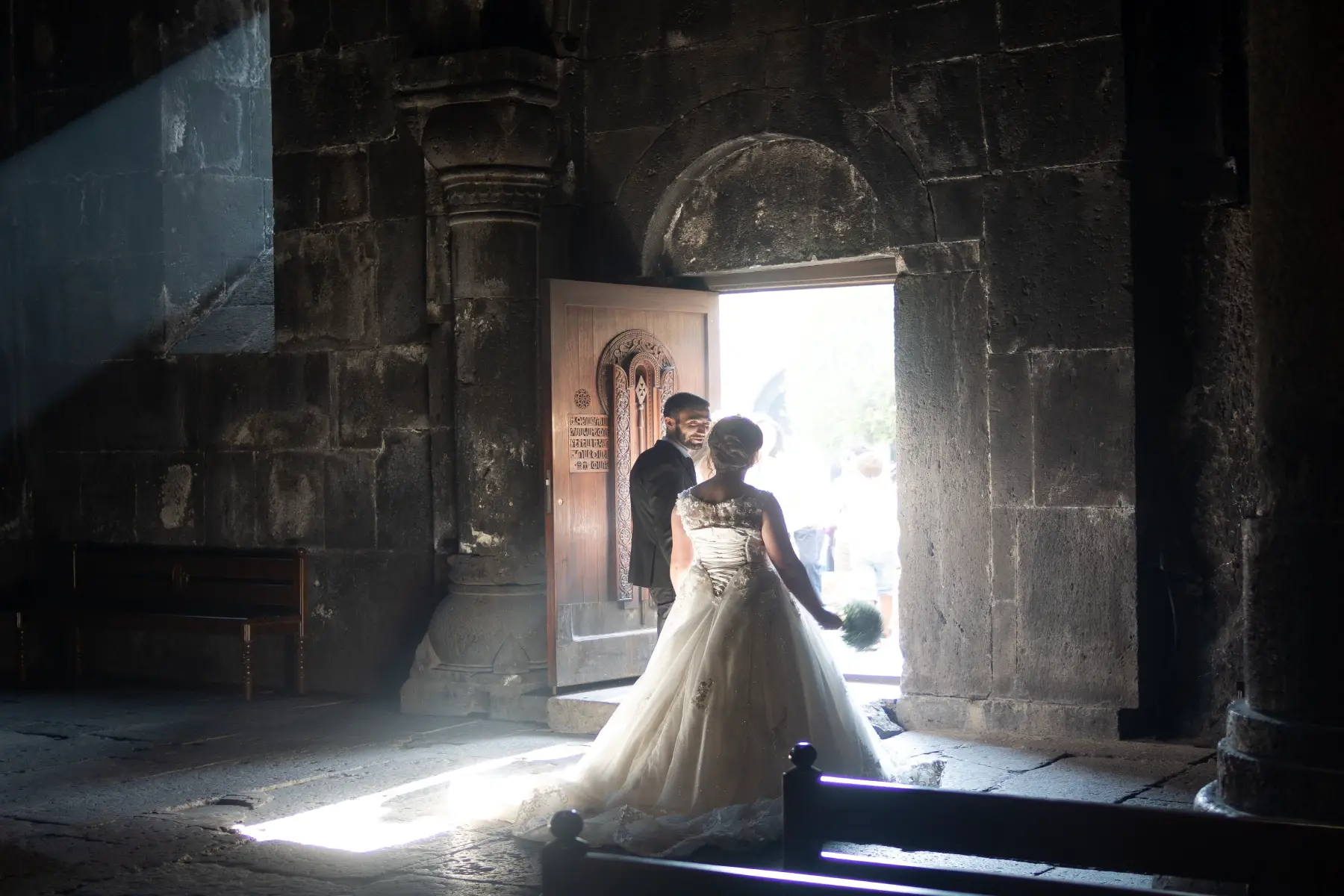 Bridal couple standing in the doorway of a historic, stone cathedral, light streaming  inside