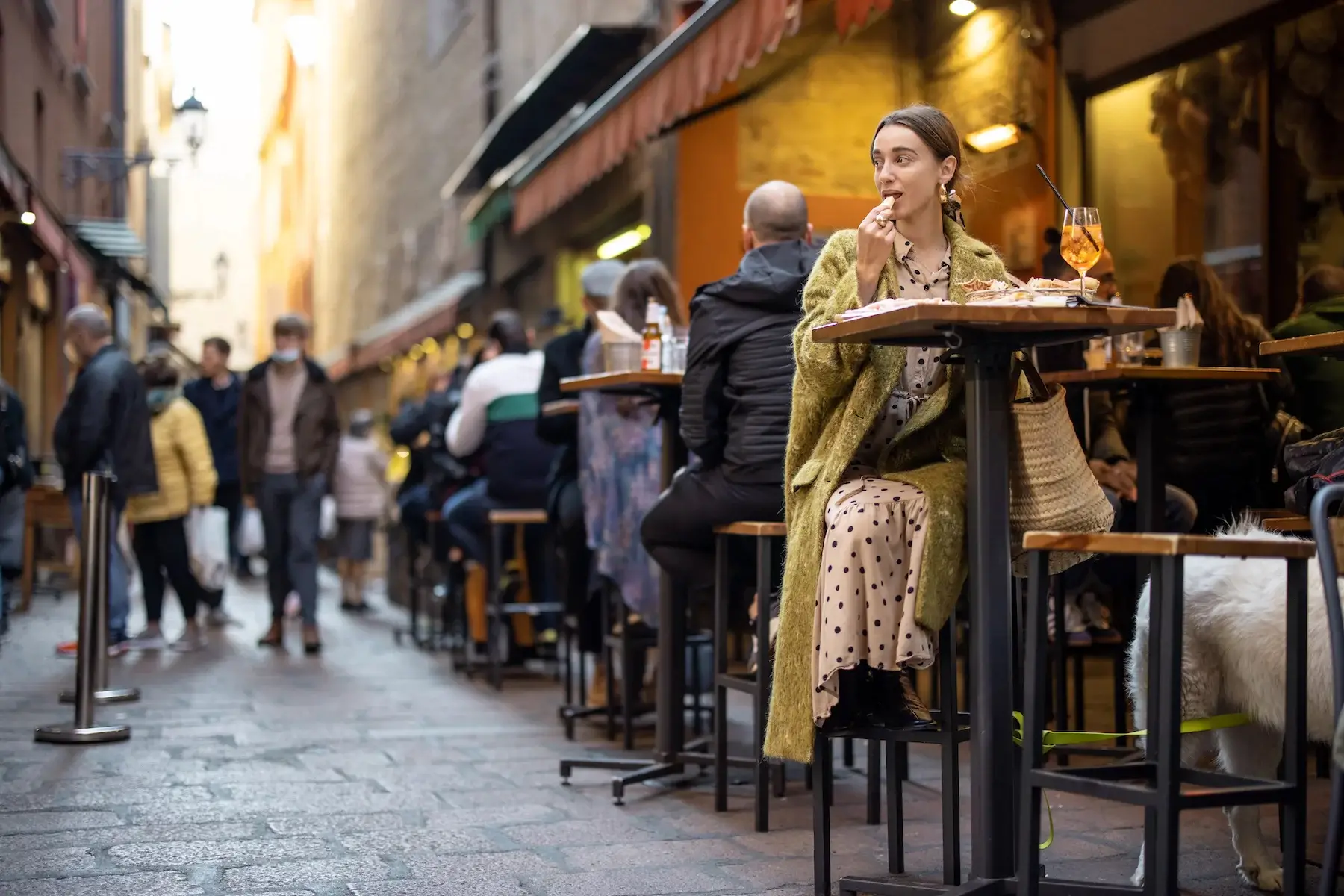 Woman sits alone with an Aperol Spritz at an outdoor bar on a narrow street in an Italian city