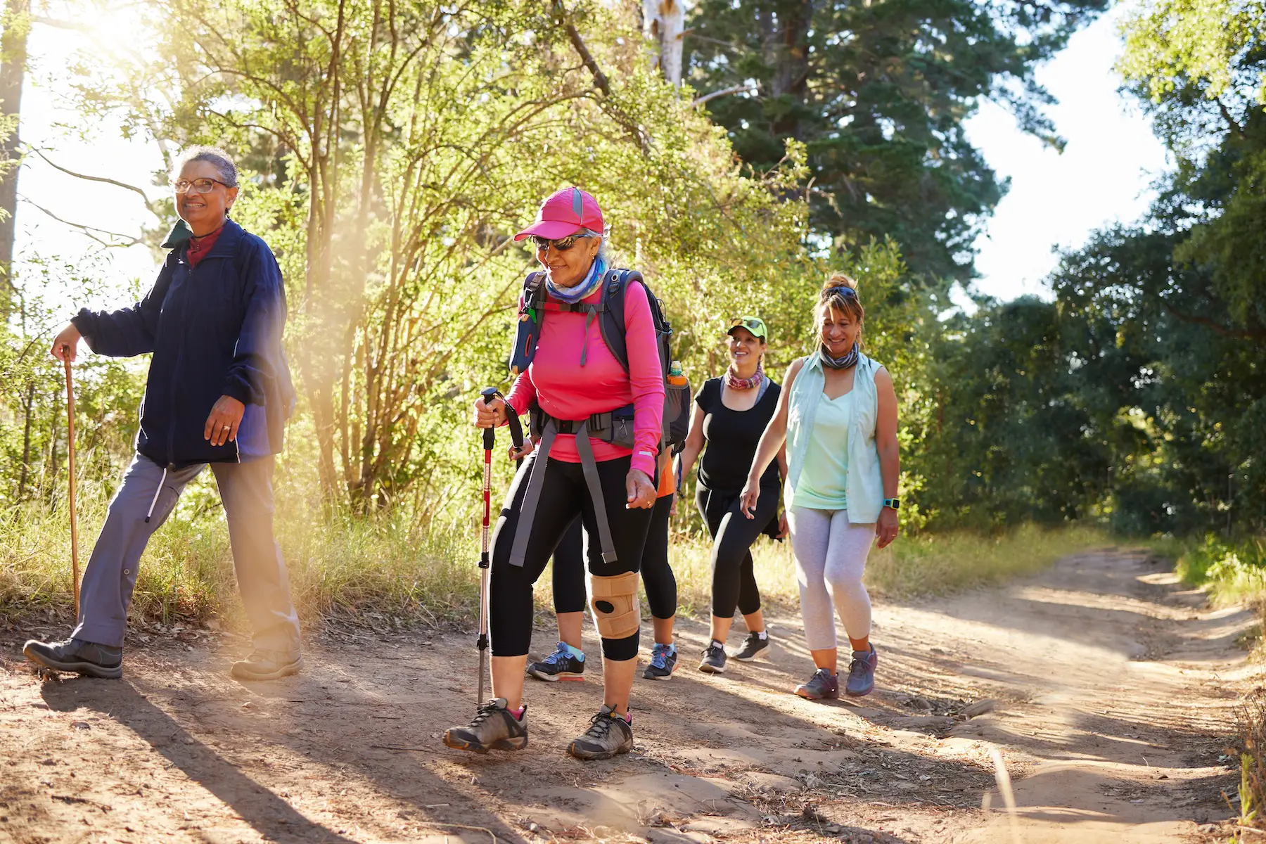 A group of five active women hike down a dirt path in a wooded area on a bright day.