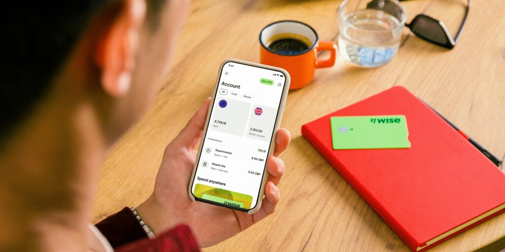 A person holding a phone with the Wise app opened. On the table there is a coffee, a notebook and the Wise card.