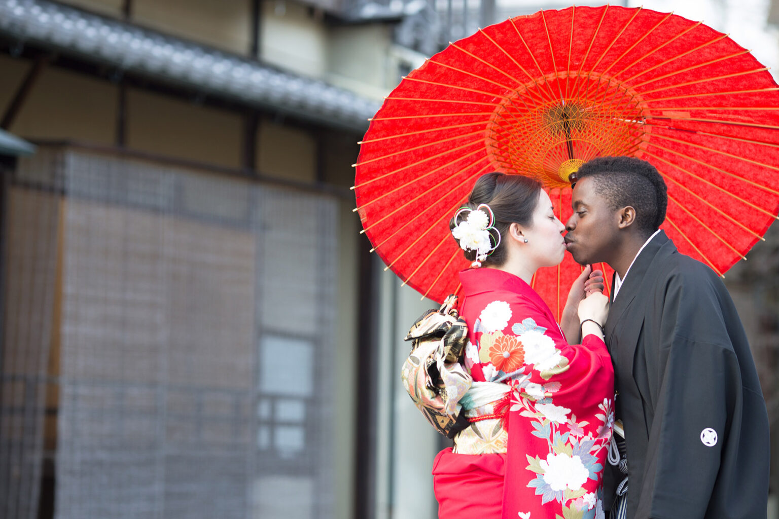 Two people kissing while standing on a red paper umbrella. They're wearing traditional red and black kimonos.