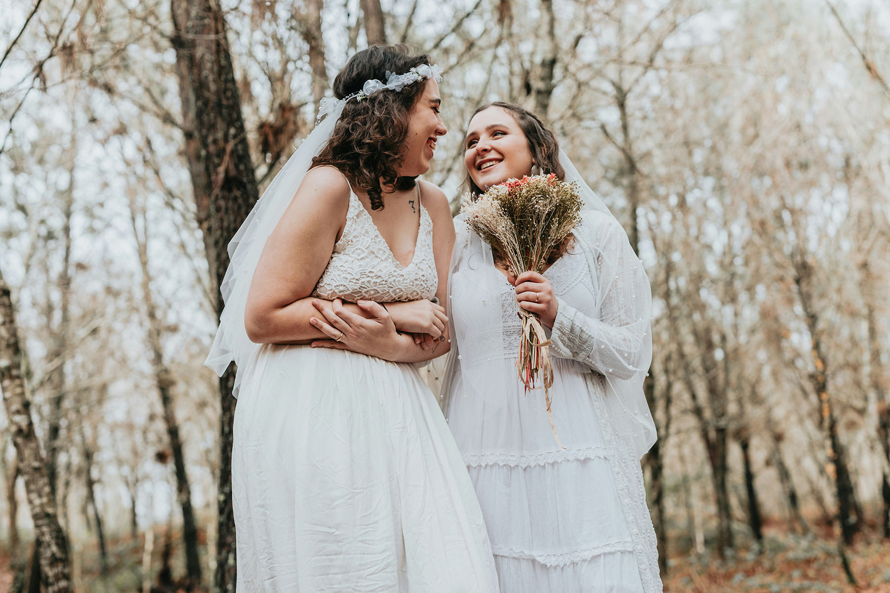 Two brides dressed in white, beaming with happiness. They're having a boho forest wedding.