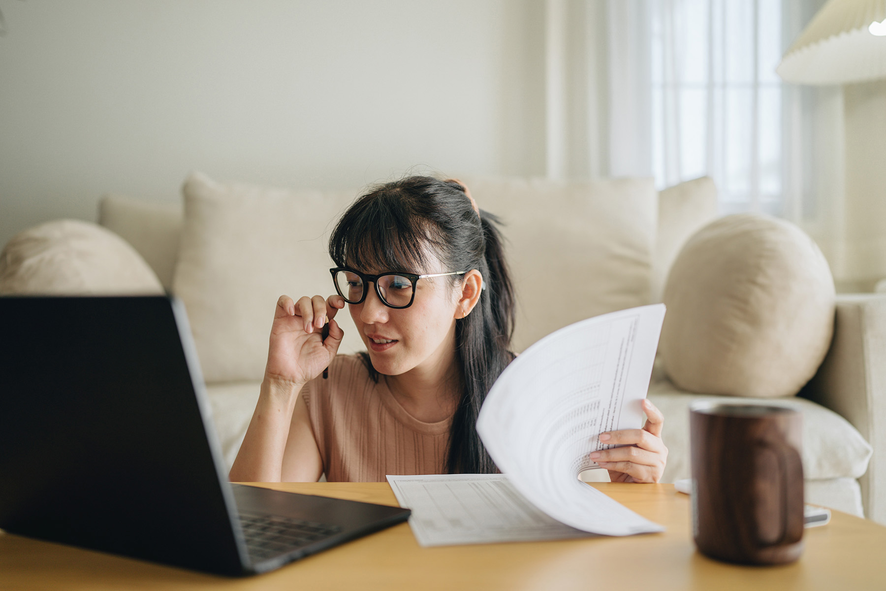 Woman sitting on the floor of her living room, near the coffee table. She's looking at a laptop, readjusting her glasses, and leaving through financial reports.
