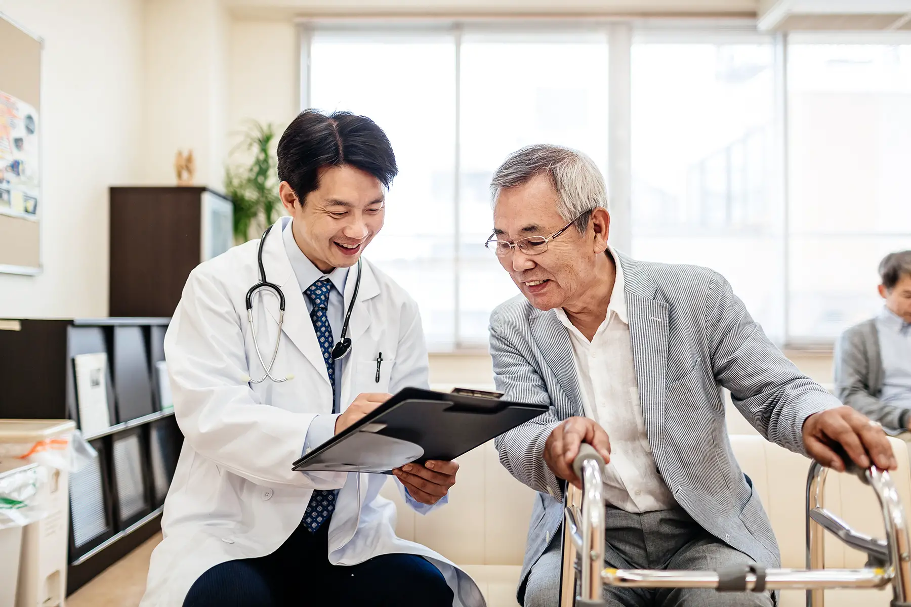 A Japanese doctor discusses his treatment plan (on a clipboard) with an older adult patient. Both are sitting and patient uses a walking frame