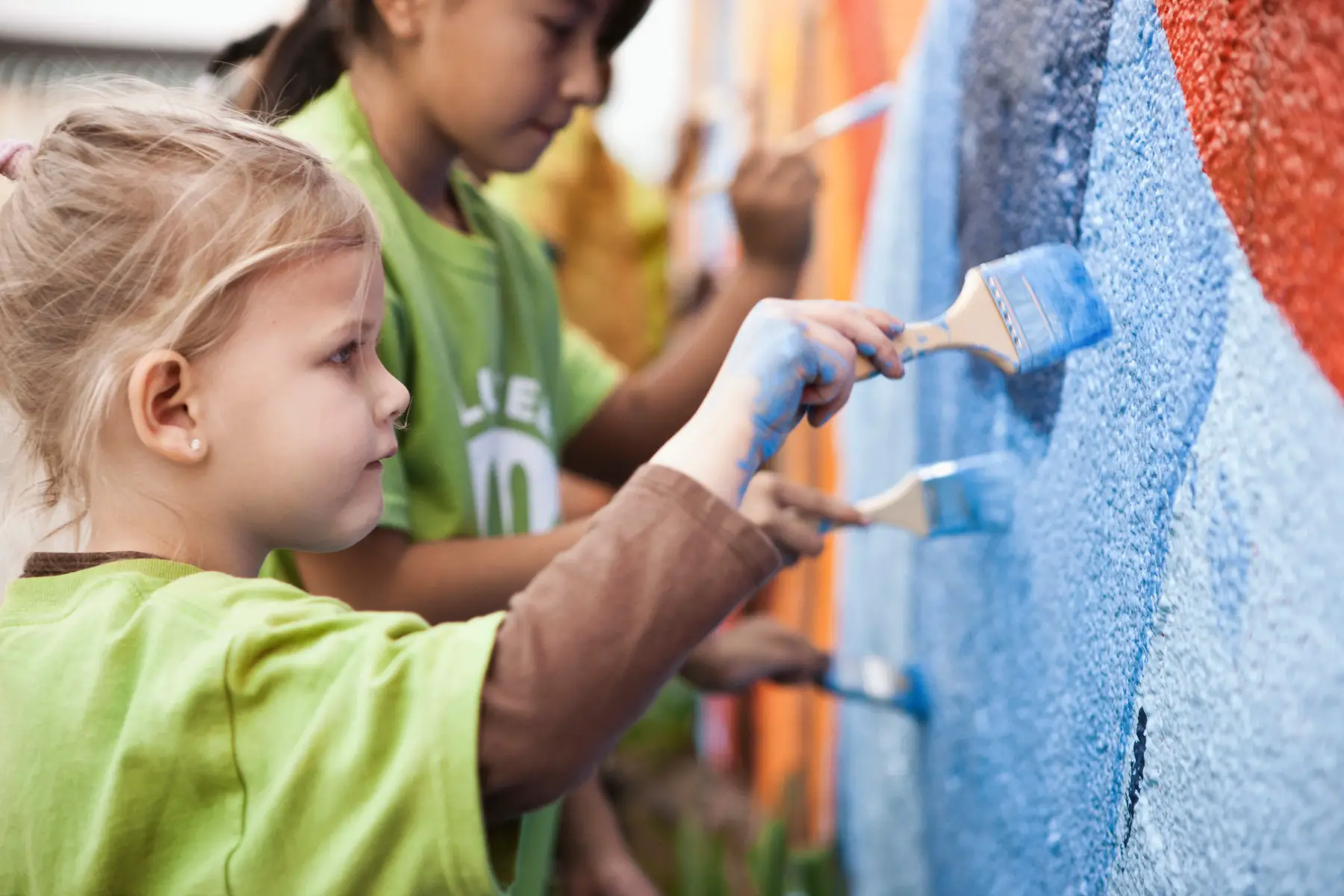 a profile shot of two little girls wearing green t-shirts and painting a wall with colorful paints