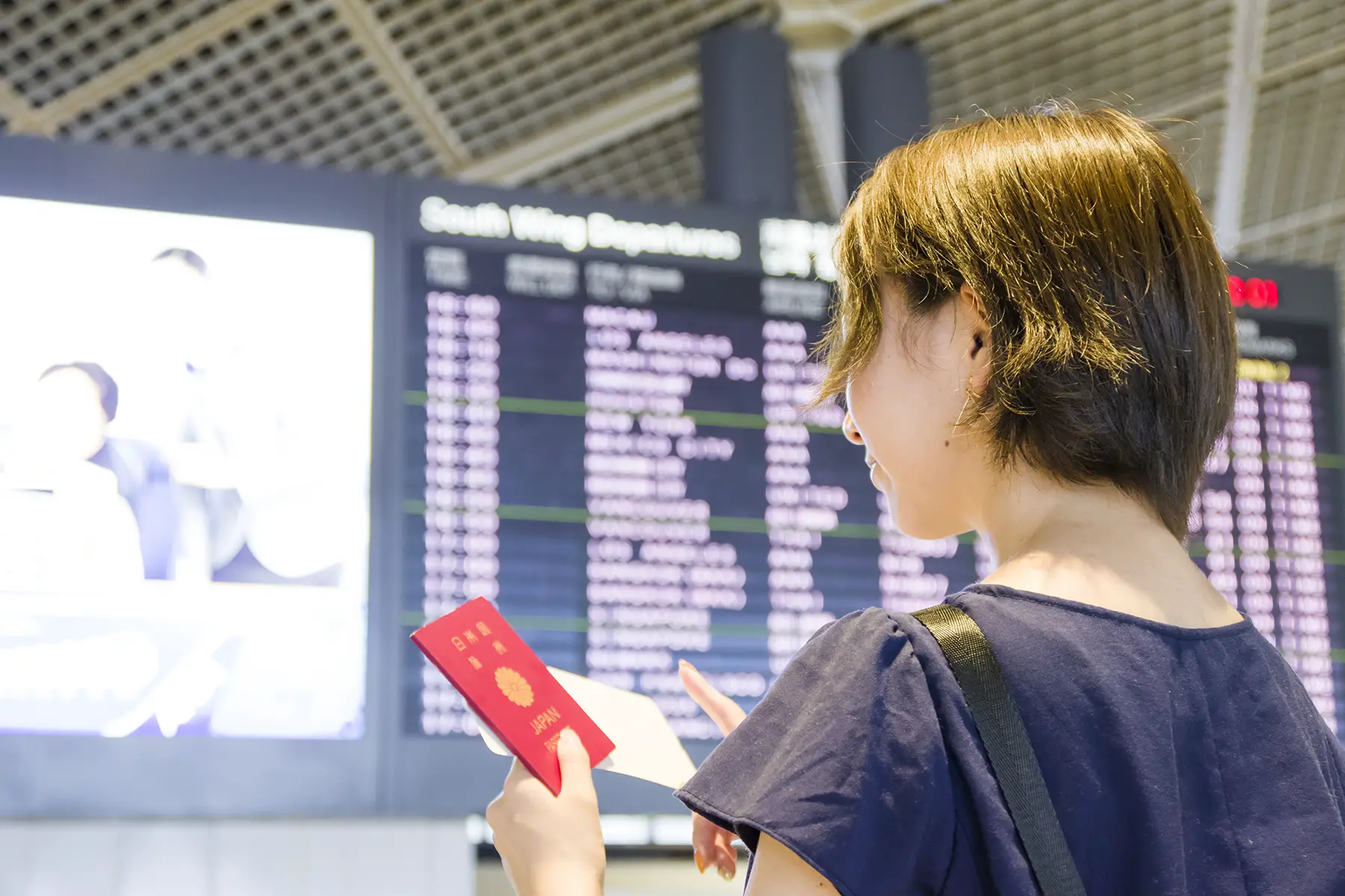 Woman with Japanese passport in hand at airport, flight status electronic boards in background