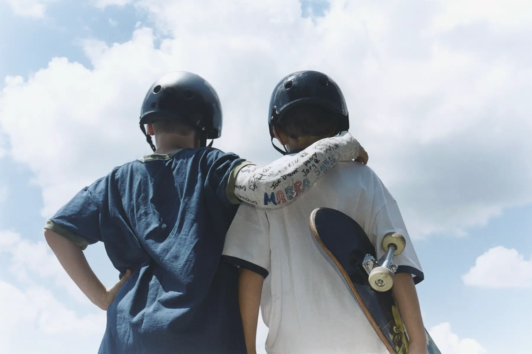 Two young skateboarders from behind with helmets, one hold a skateboard and other has his arm in a cast around the friend's shoulders