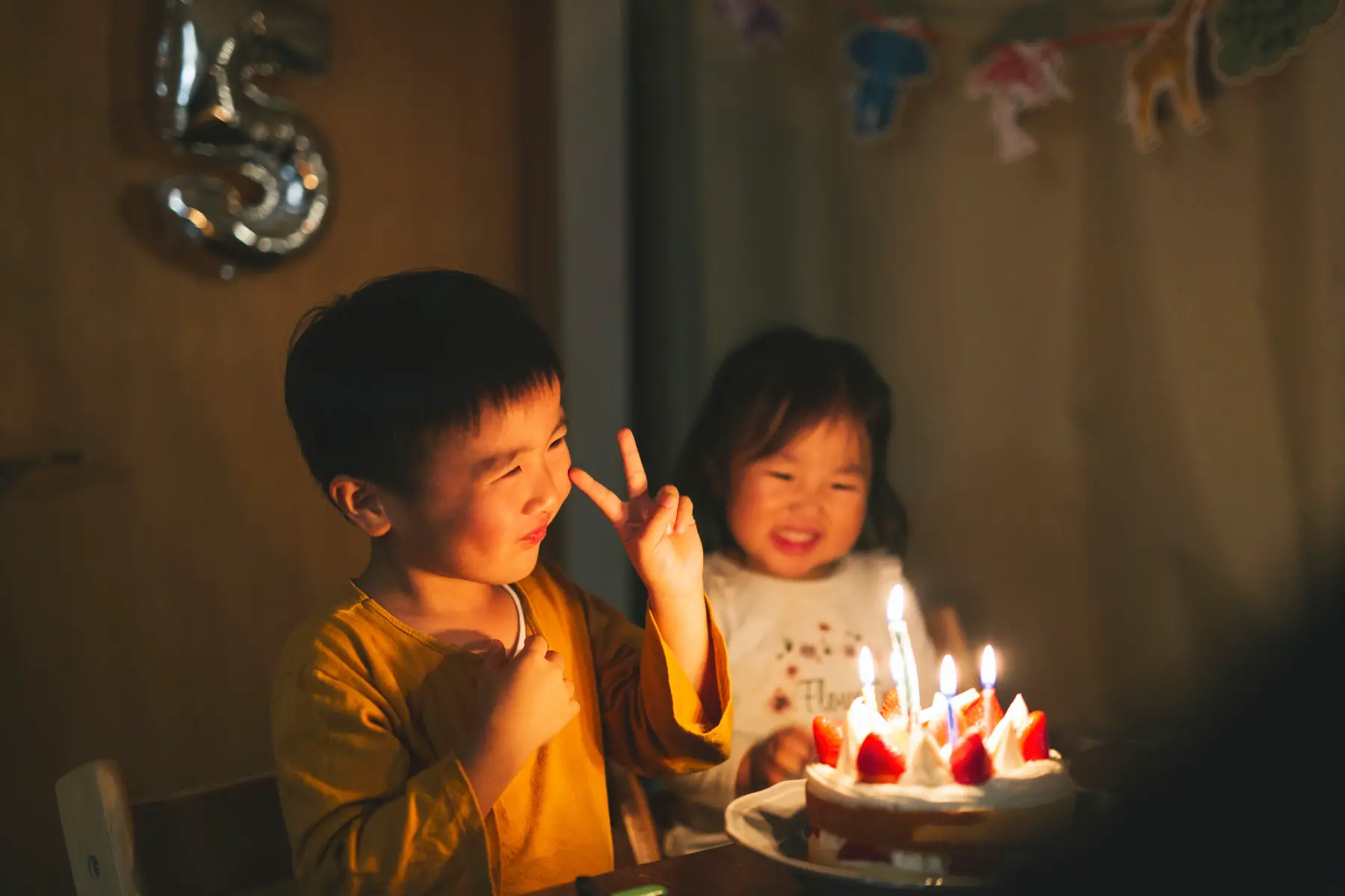 Child holds up peace next to his sister as they sit in front of a lit birthday cake