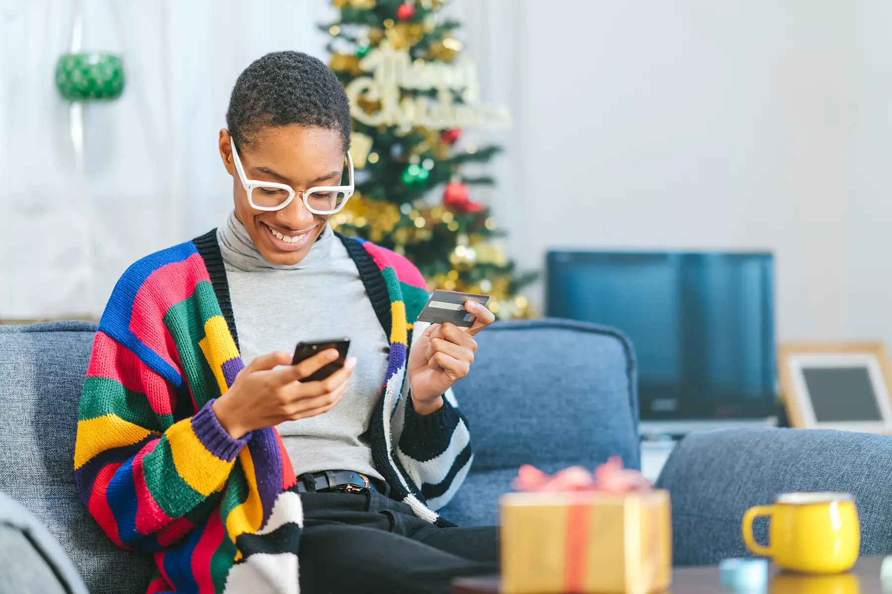 A woman in a brightly colored stripy jumper sits on the sofa, paying for something on her phone.