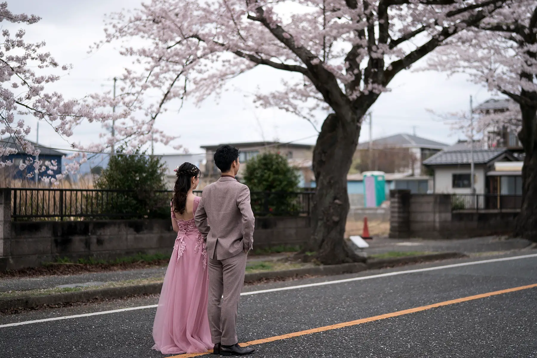 A couple in a grey suit and a pink dress standing for photos under a cherry blossom tree.