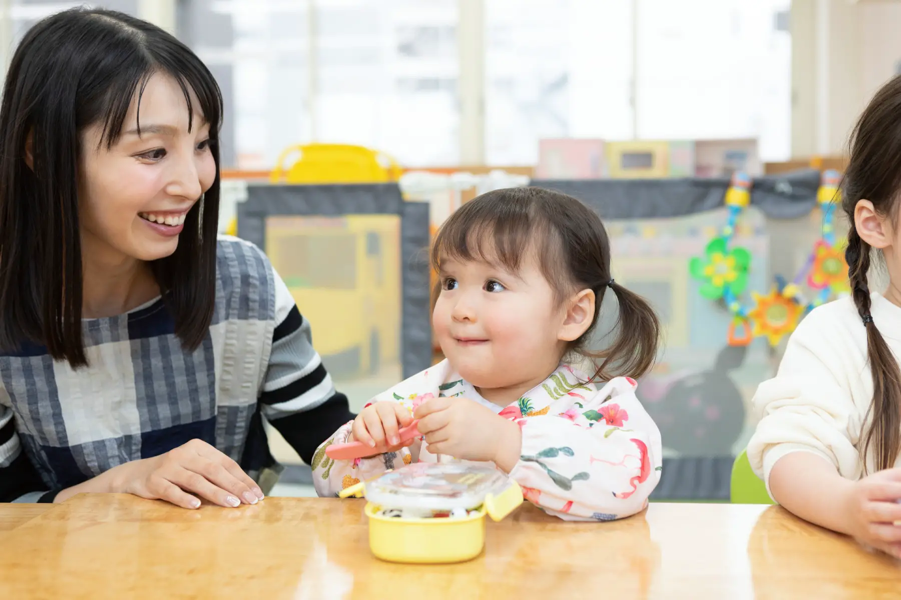 a little Japanese girl sat next to her daycare leader enjoying lunch at her desk 