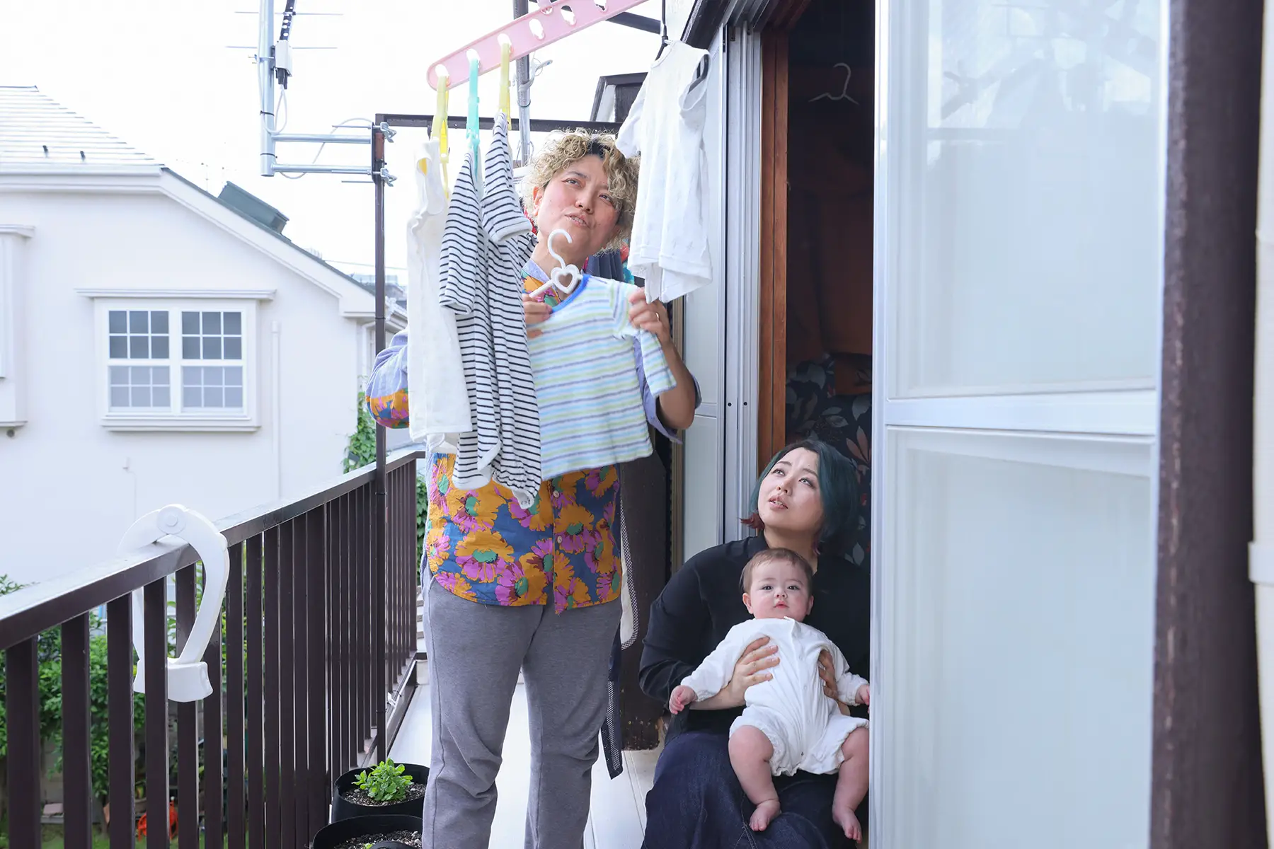 On a balcony, one mom hangs up the washing, while the other mom holds their baby