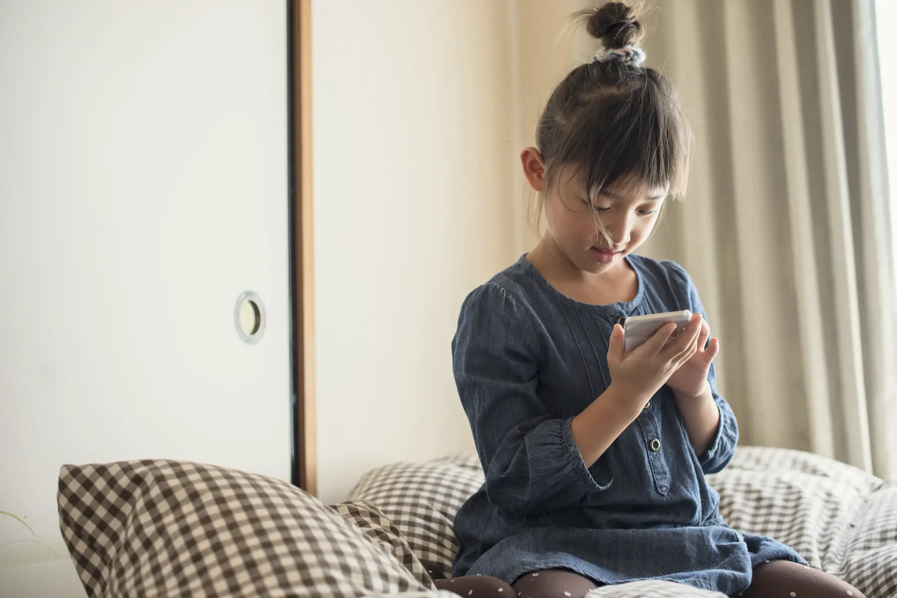 A little girl with bangs and hair in a bun sits on a bed playing with a smartphone