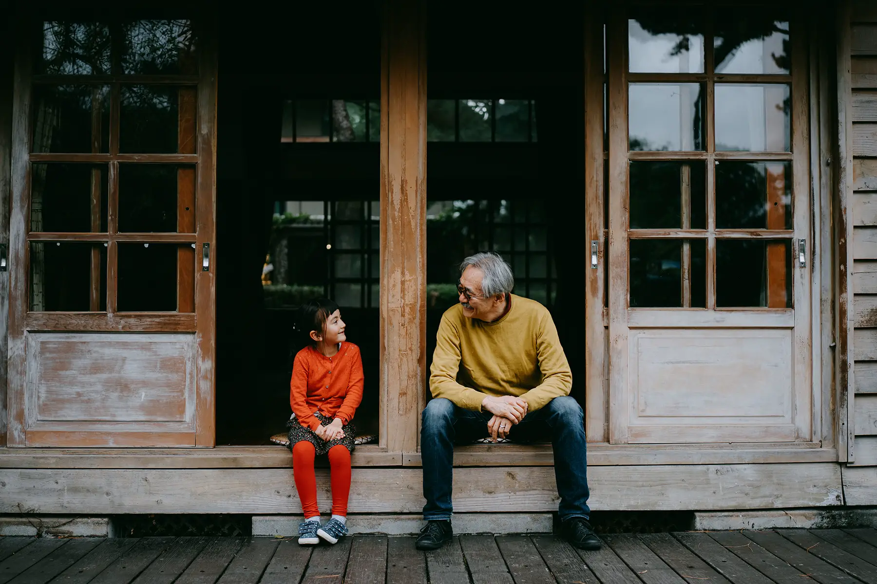 A granddaughter and a grandfather sit on a porch together, smiling at each other.