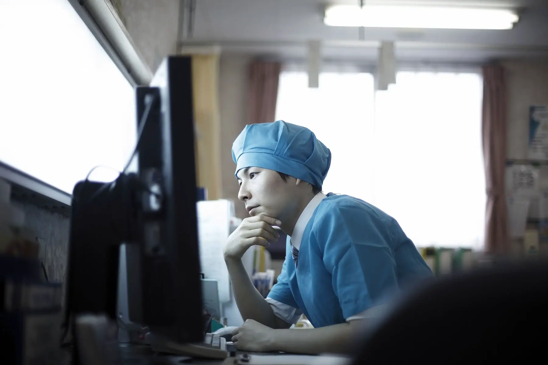 A doctor in scrubs sits looking at his desktop computer screen, deep in thought with his hand on his chin