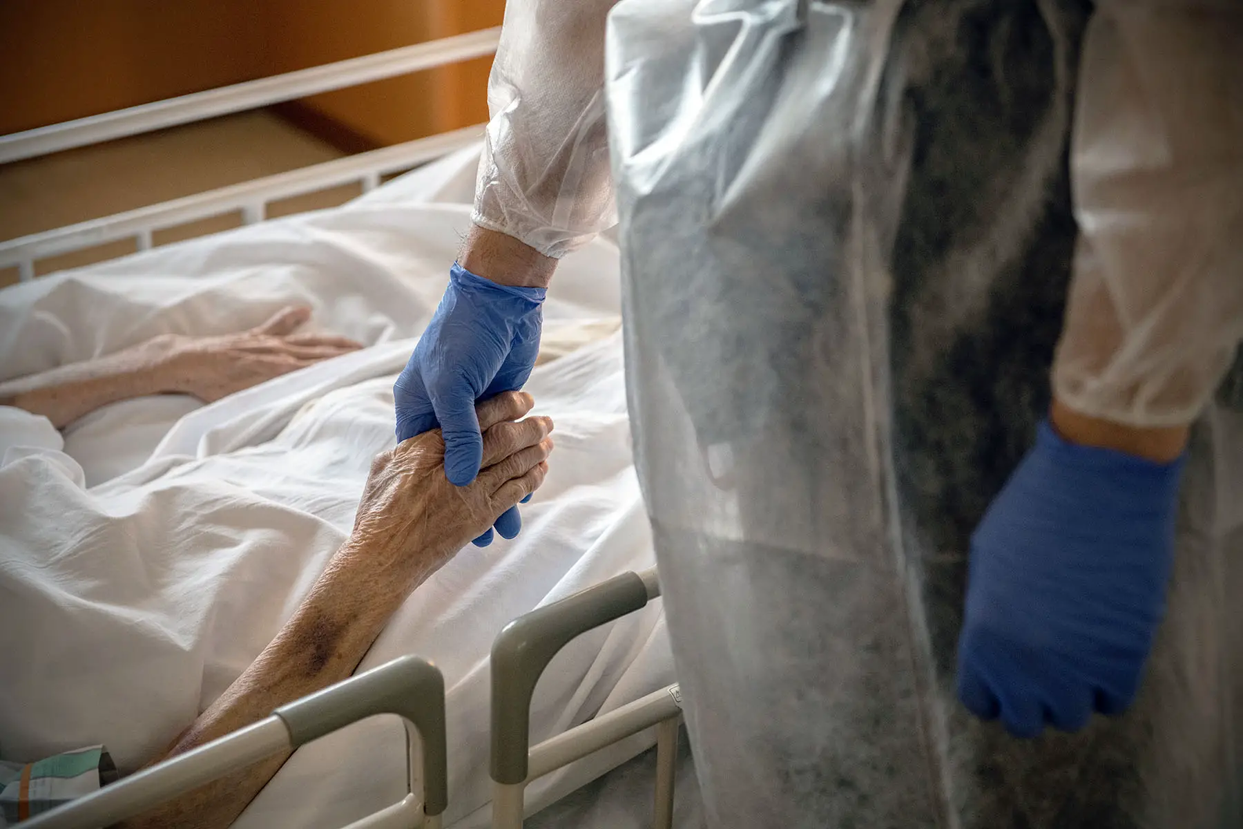 Old patient holding the gloved hand of a person standing by.